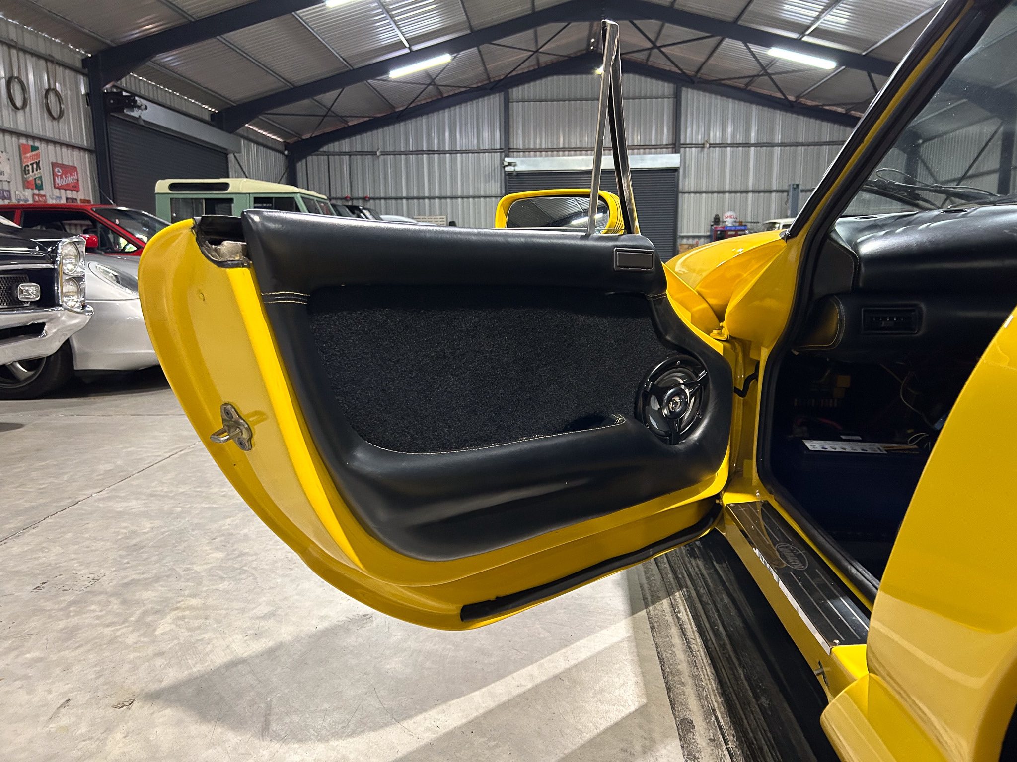 Open yellow car door in a garage filled with classic cars, showing black interior trim and chrome accents.