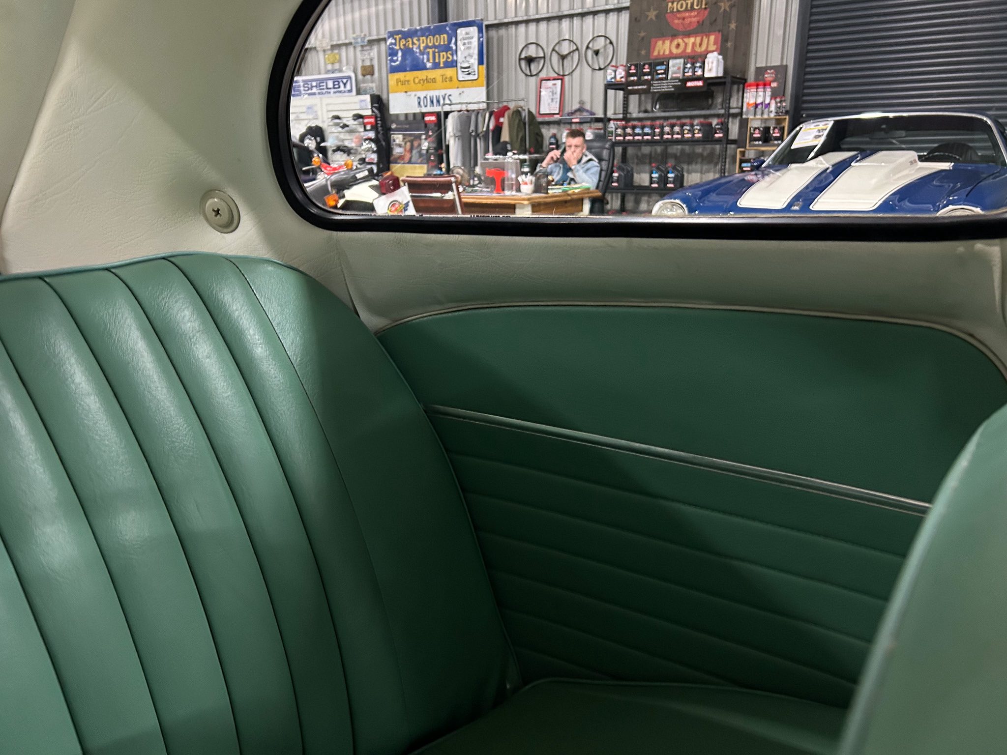 Interior of a vintage car with green upholstery, looking out at a workshop with a blue-and-white classic car and a man at a desk.