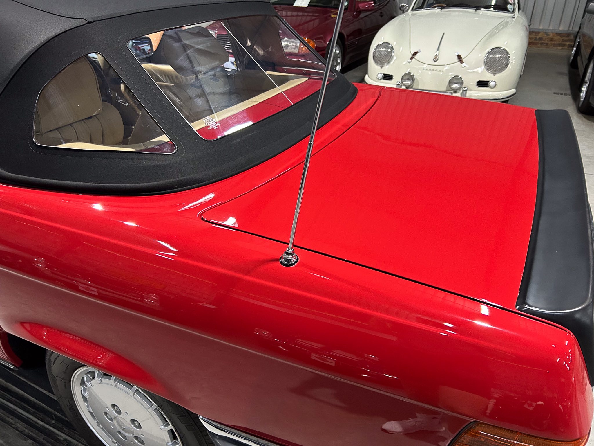 Red vintage convertible in a showroom with a black soft top and chrome antenna, other classic cars visible in the background