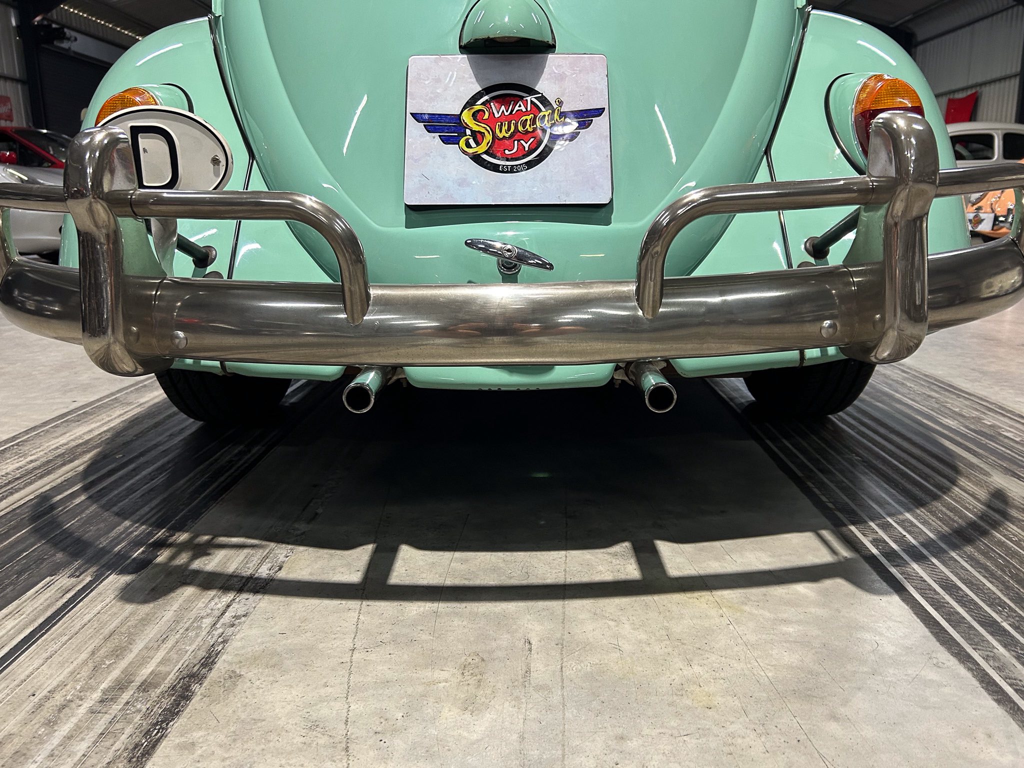 Rear view of a mint-green vintage car with a heavy chrome bumper and a square license plate featuring a circular logo (EST 2015).