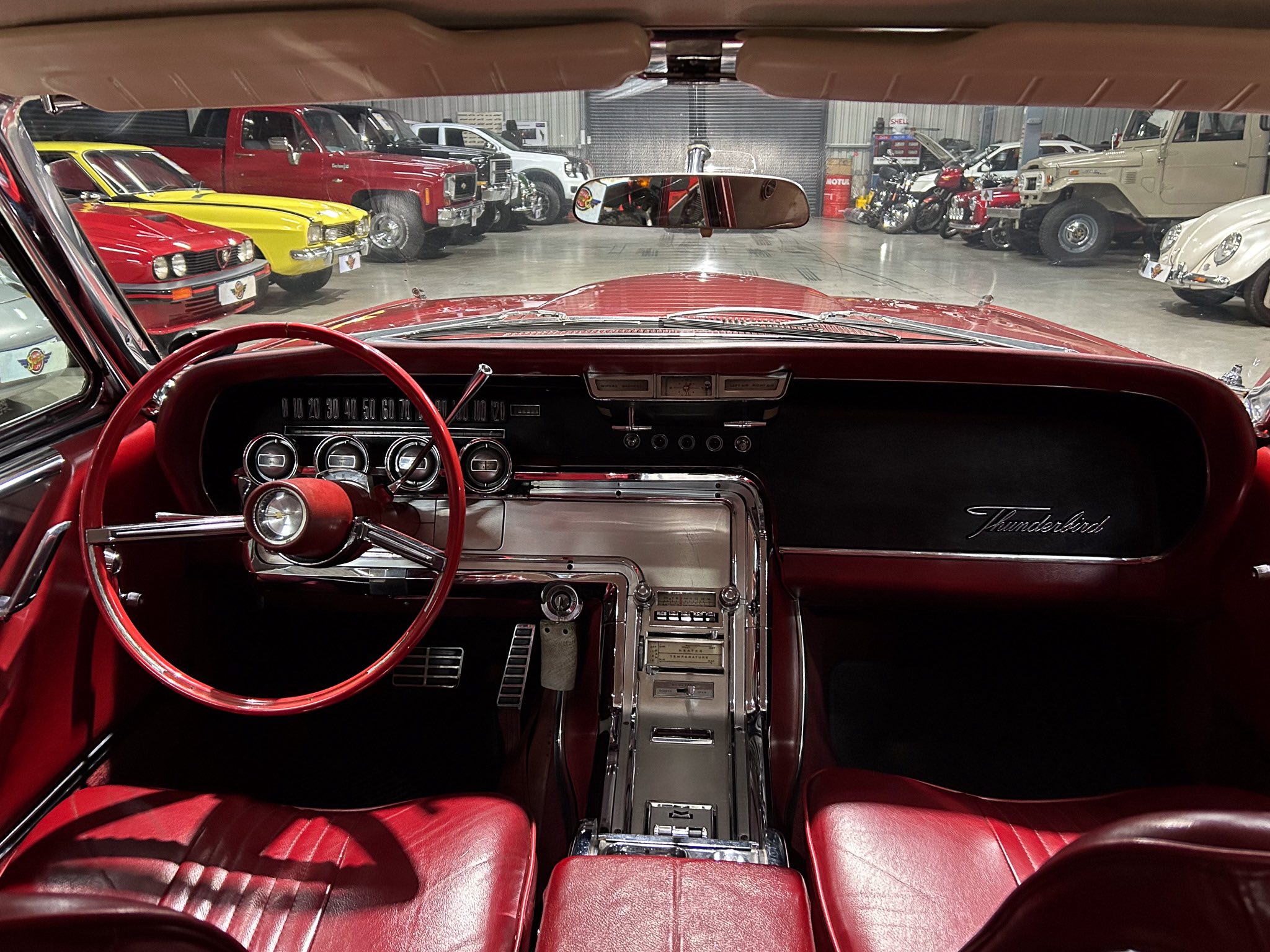 View from the driver’s seat of a red vintage car, overlooking a showroom filled with classic cars and motorcycles in the background.