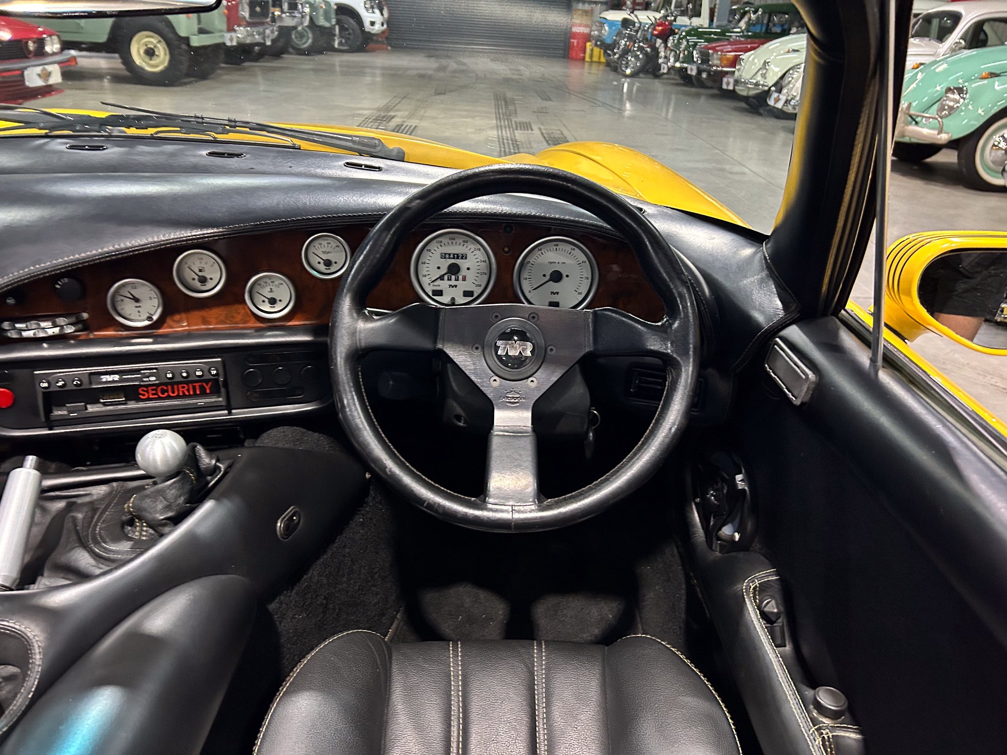 Driver's view of a yellow vintage car interior with a black dashboard, round gauges, and a gear shifter; other classic cars are visible in a showroom.
