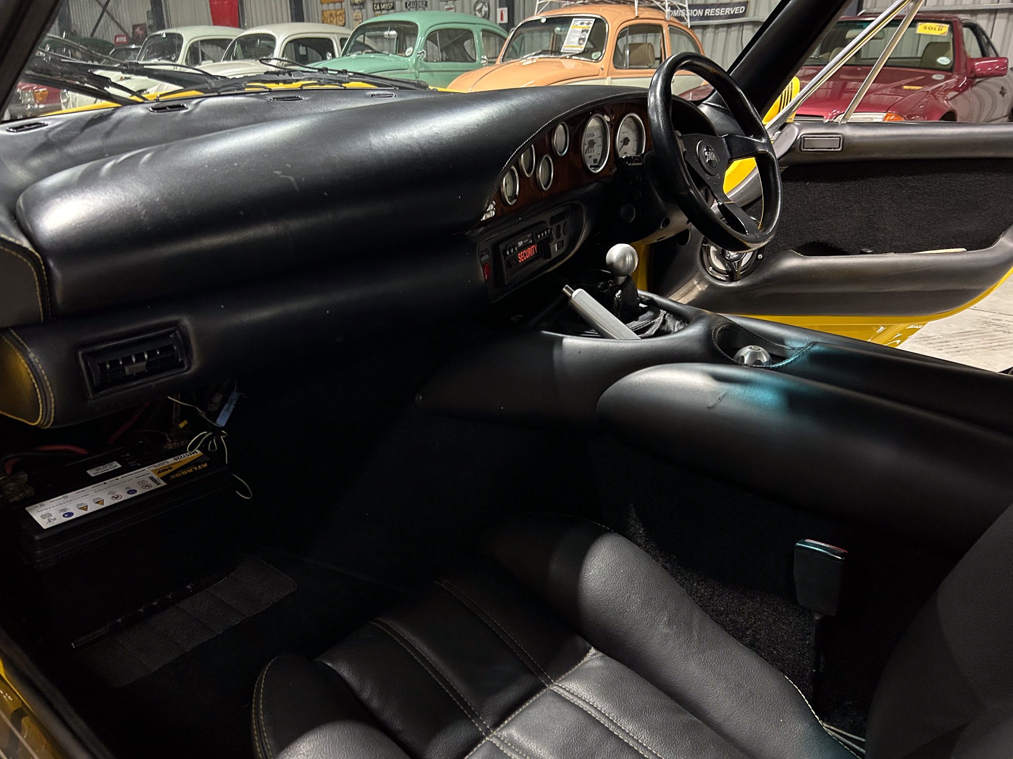 Interior view of a classic car: black leather dashboard, steering wheel, and instruments; showroom with vintage cars visible outside