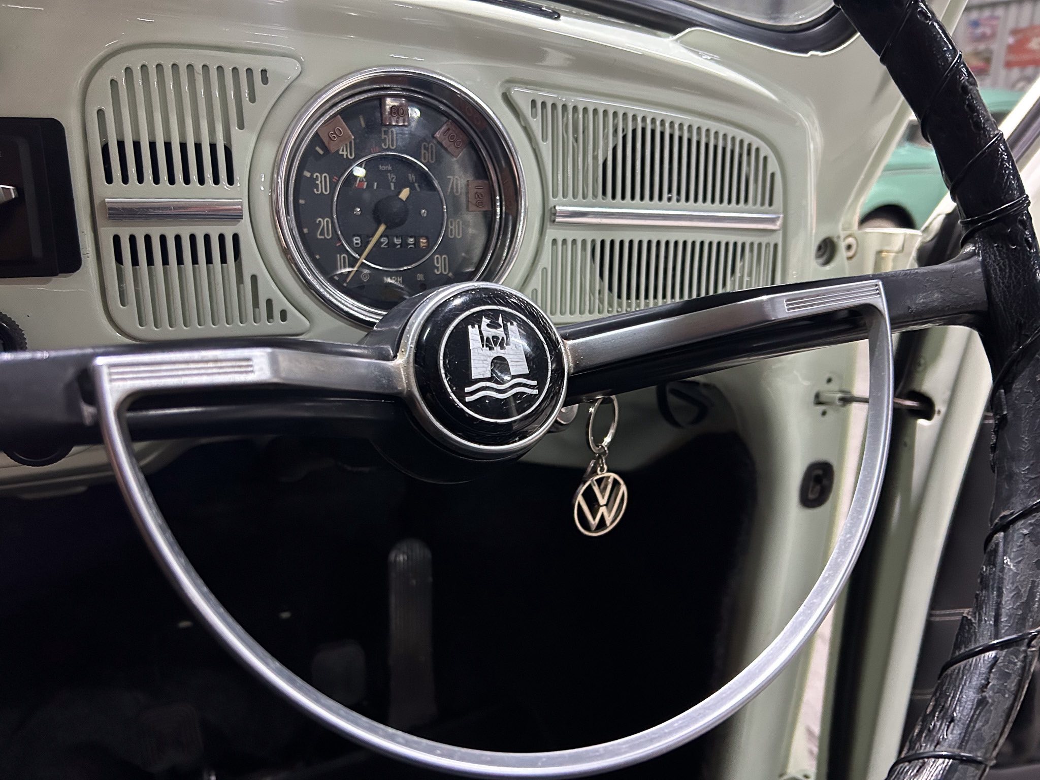 Vintage car dashboard with a circular speedometer and VW steering wheel emblem visible in the center, greenish interior panels surrounding the gauges.