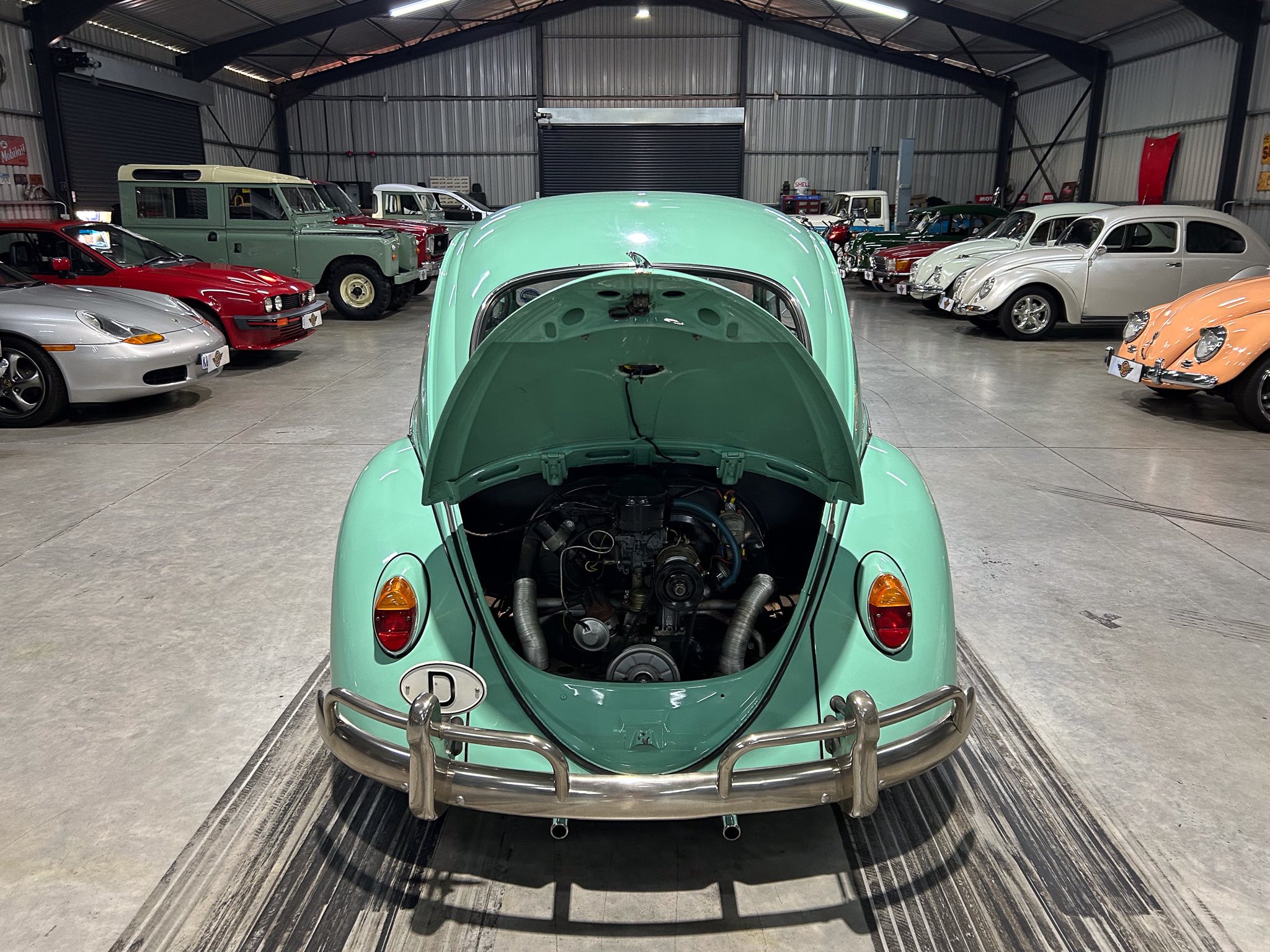 Interior of a vintage car showroom with a mint-green car in the foreground and rows of classic cars behind it on both sides, under a metal warehouse roof.