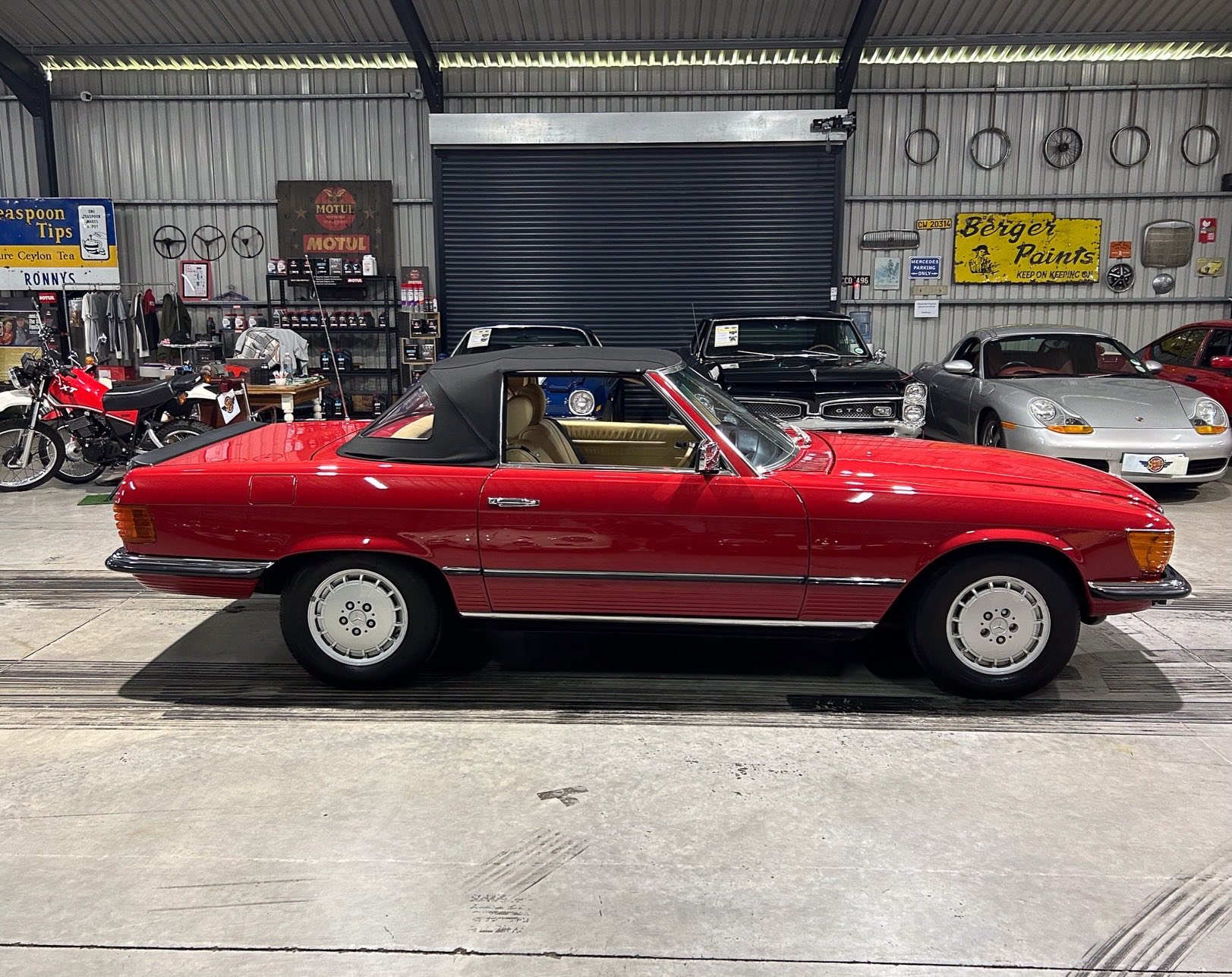Red classic convertible with black soft top inside a vintage car showroom, surrounded by other classic cars and bikes in the background.
