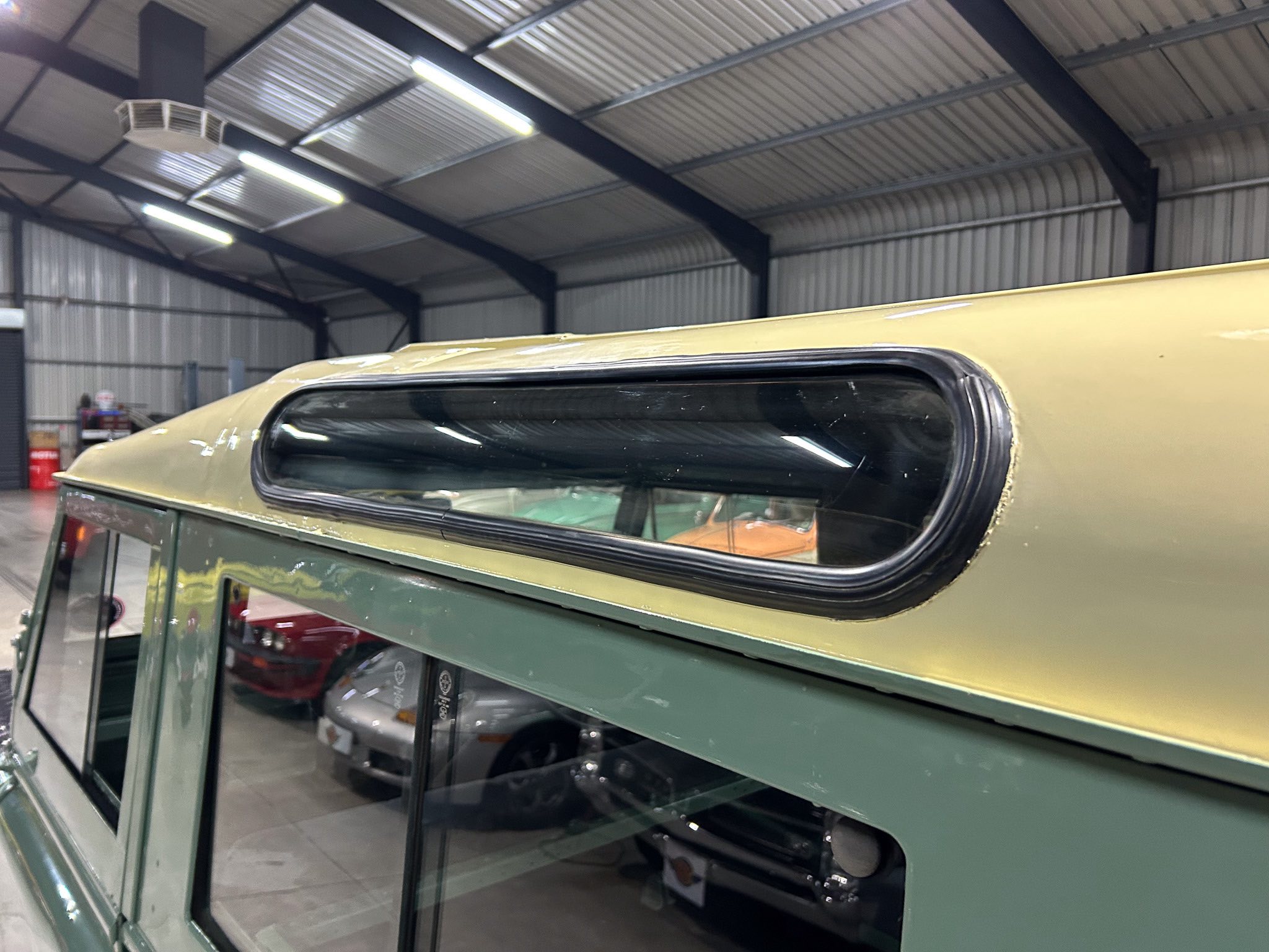 Close-up of a vintage beige roofline and green body of a classic car inside a workshop with metal beams and fluorescent lights above.