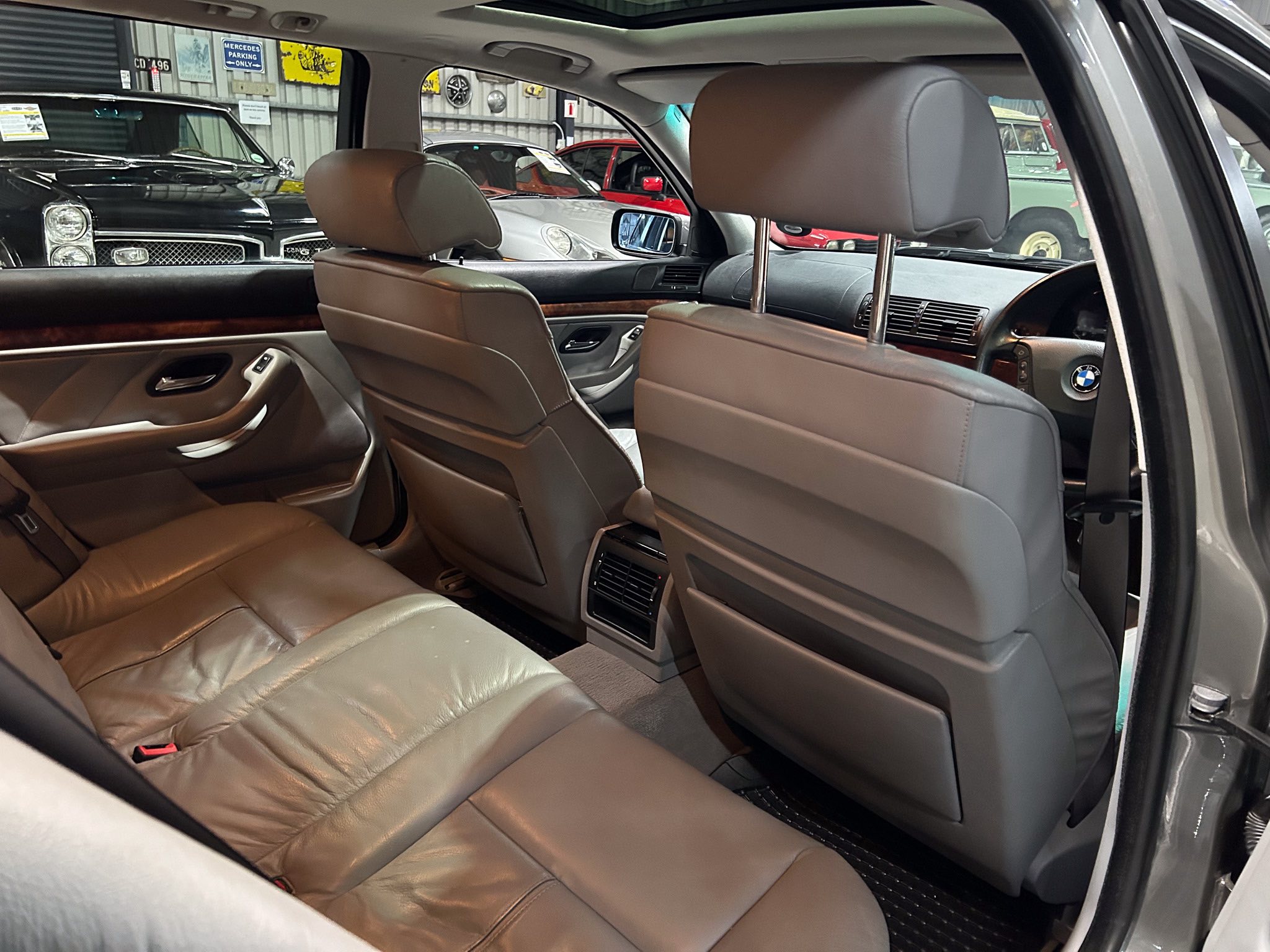 Backseat view of a tan leather car interior with front seats and center console, taken from the open rear door in a showroom filled with other vehicles outside the window
