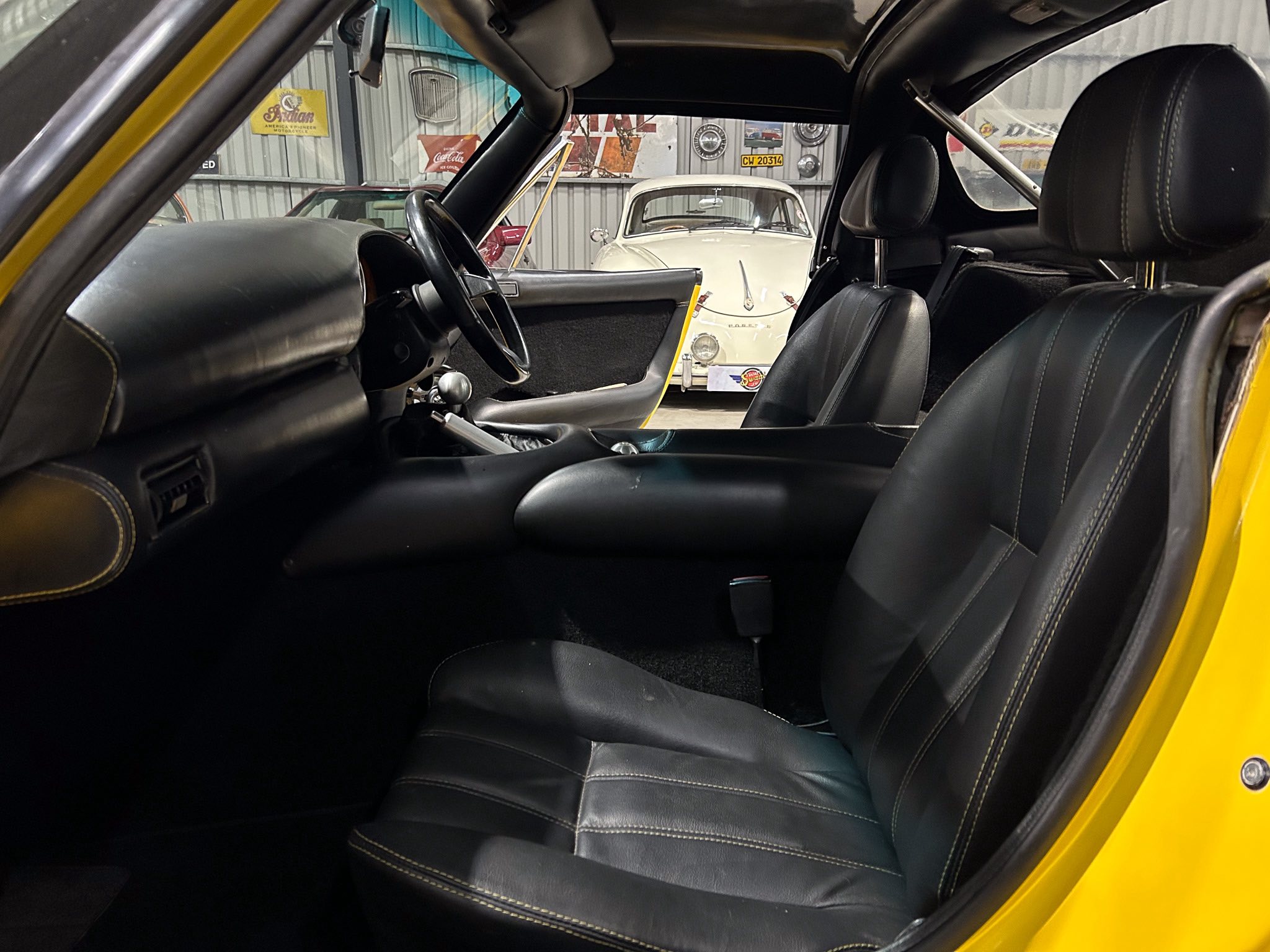 Inside a black leather car cabin, viewed toward a white vintage car parked in a workshop.