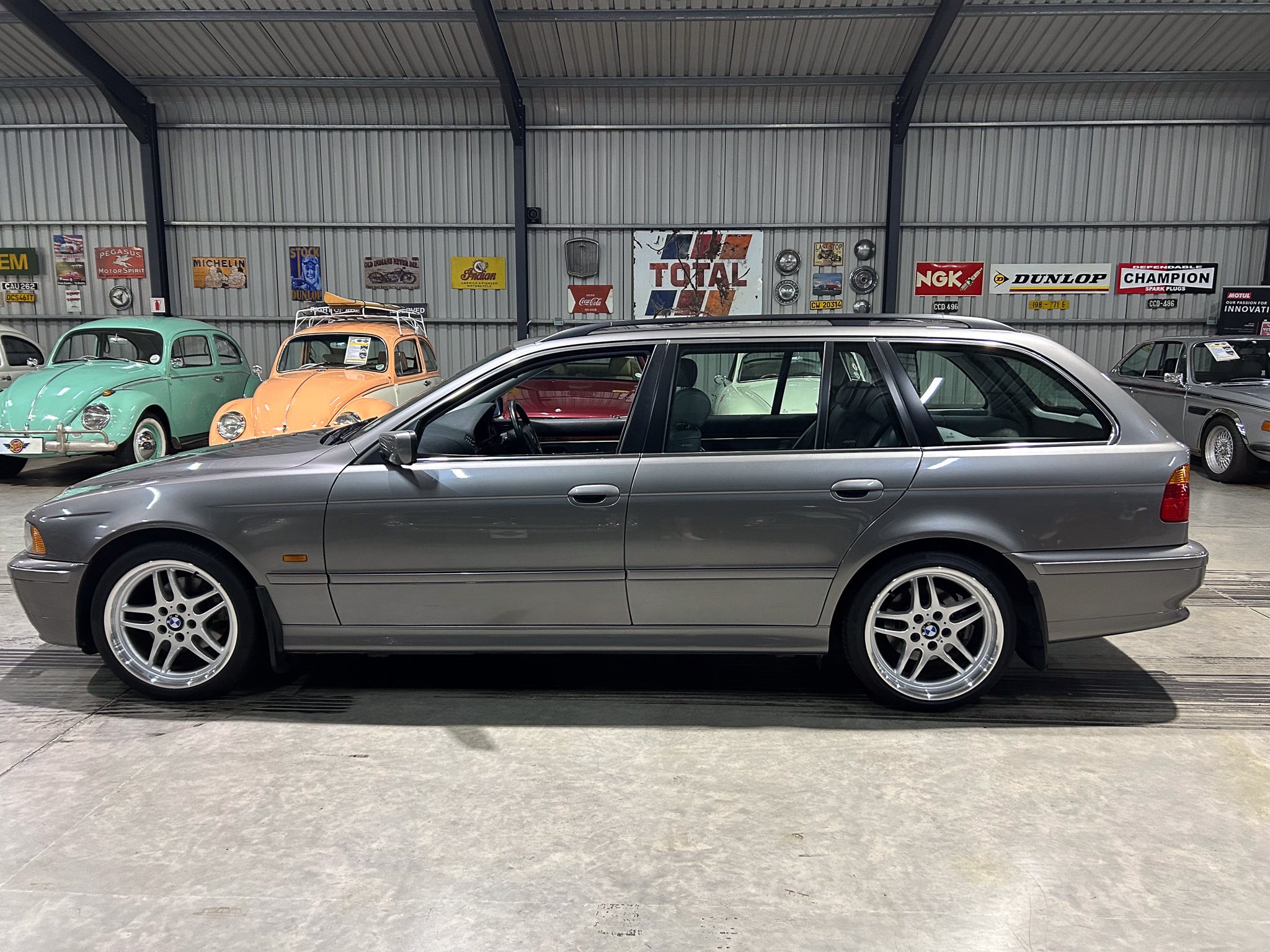 Silver BMW wagon in a retro car showroom with pastel mint and peach VW Beetles and vintage posters on a metal wall.