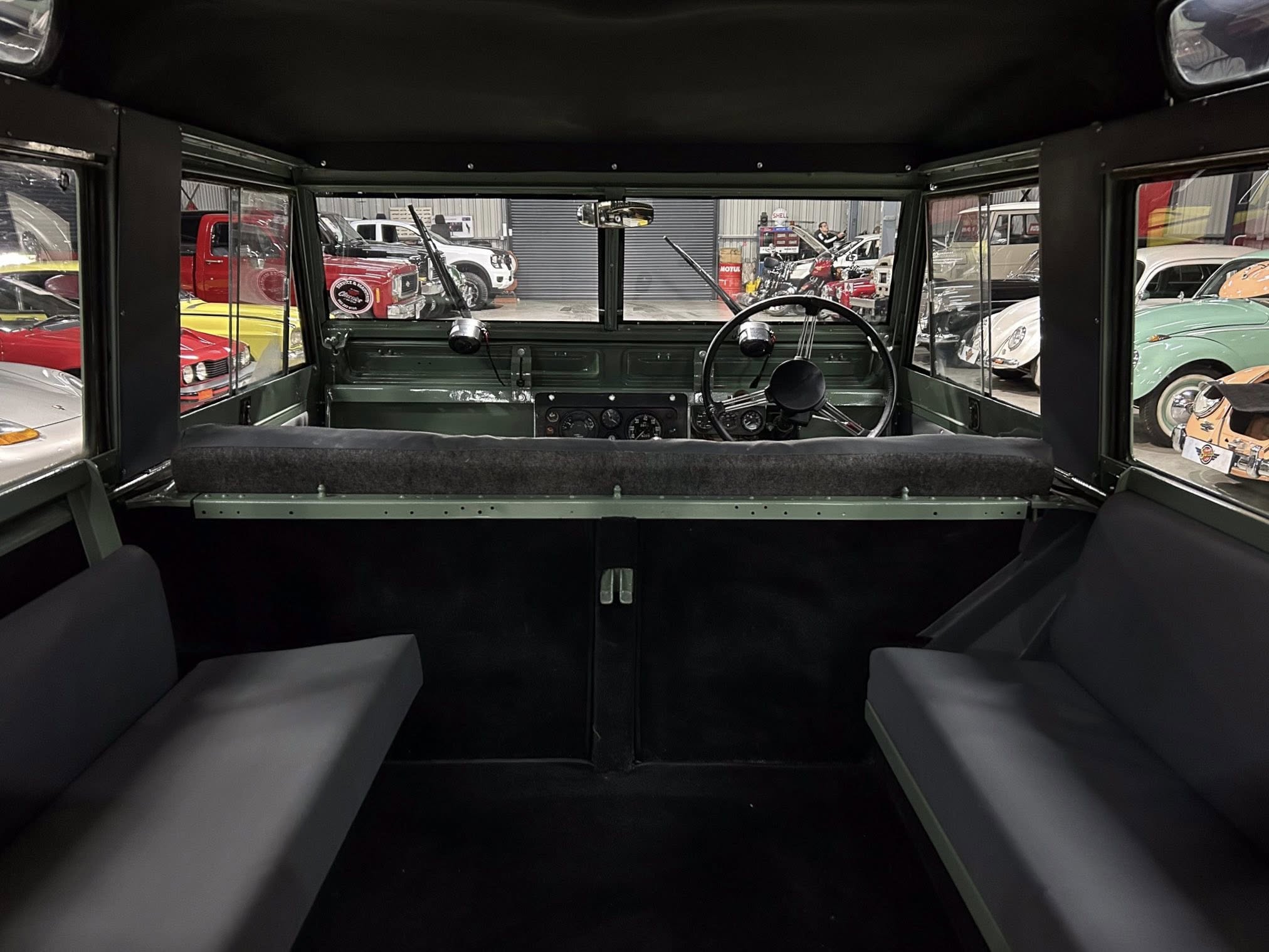 Interior view from a vintage vehicle's cabin looking forward at a showroom full of classic cars outside the windshield.
