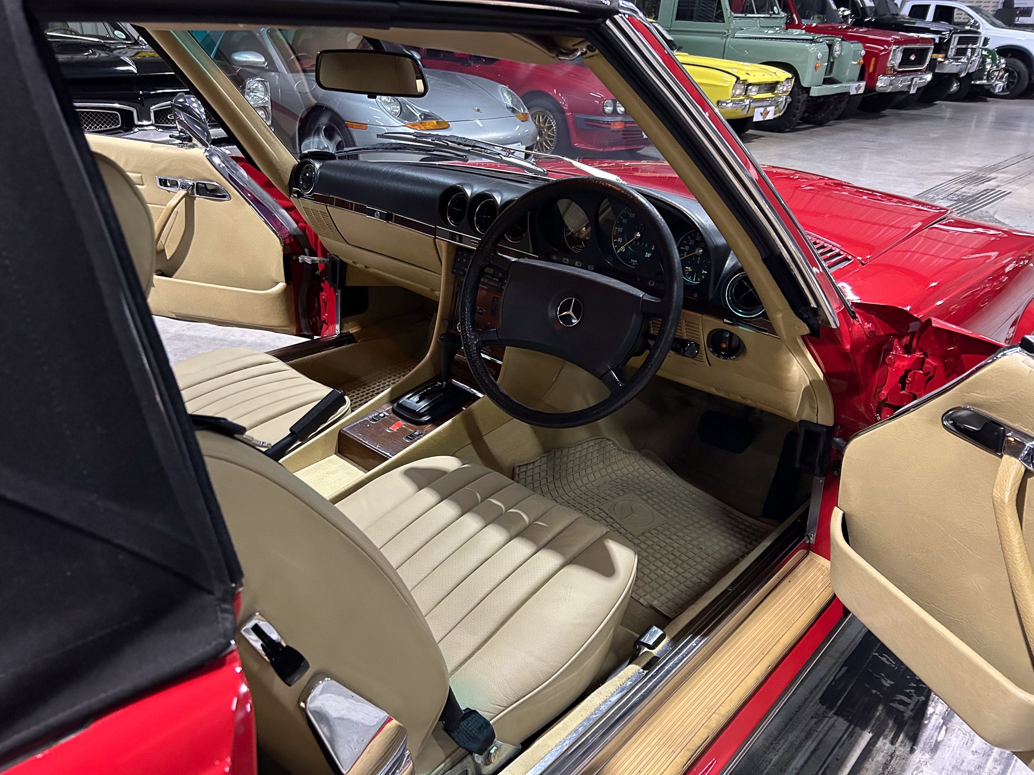 Front interior view of a red vintage Mercedes with tan leather seats, wood trim, and a black dashboard inside a car showroom.