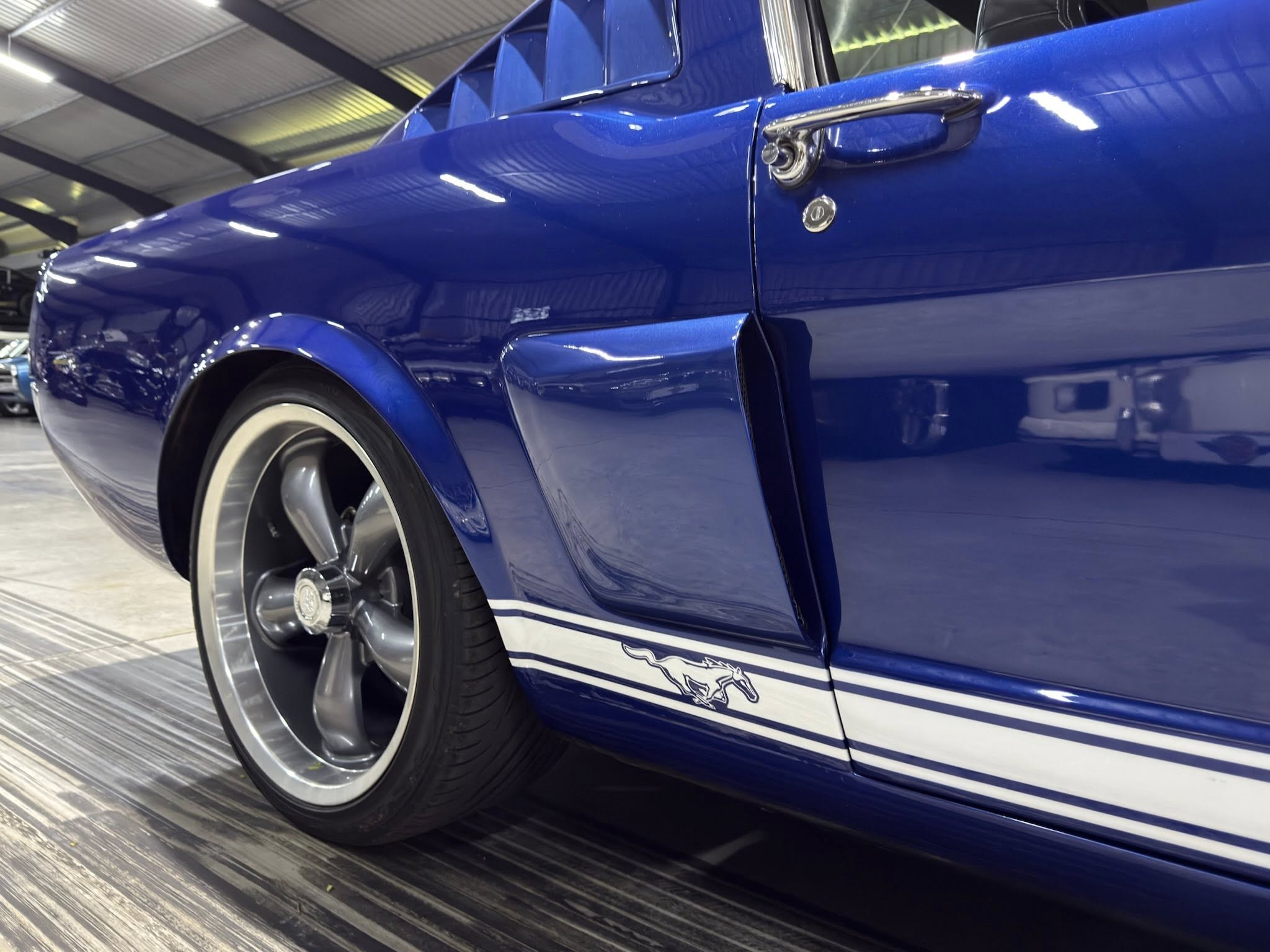 Close-up of a glossy blue vintage car door with chrome handle, running horse logo, and a visible wheel in a showroom setting.