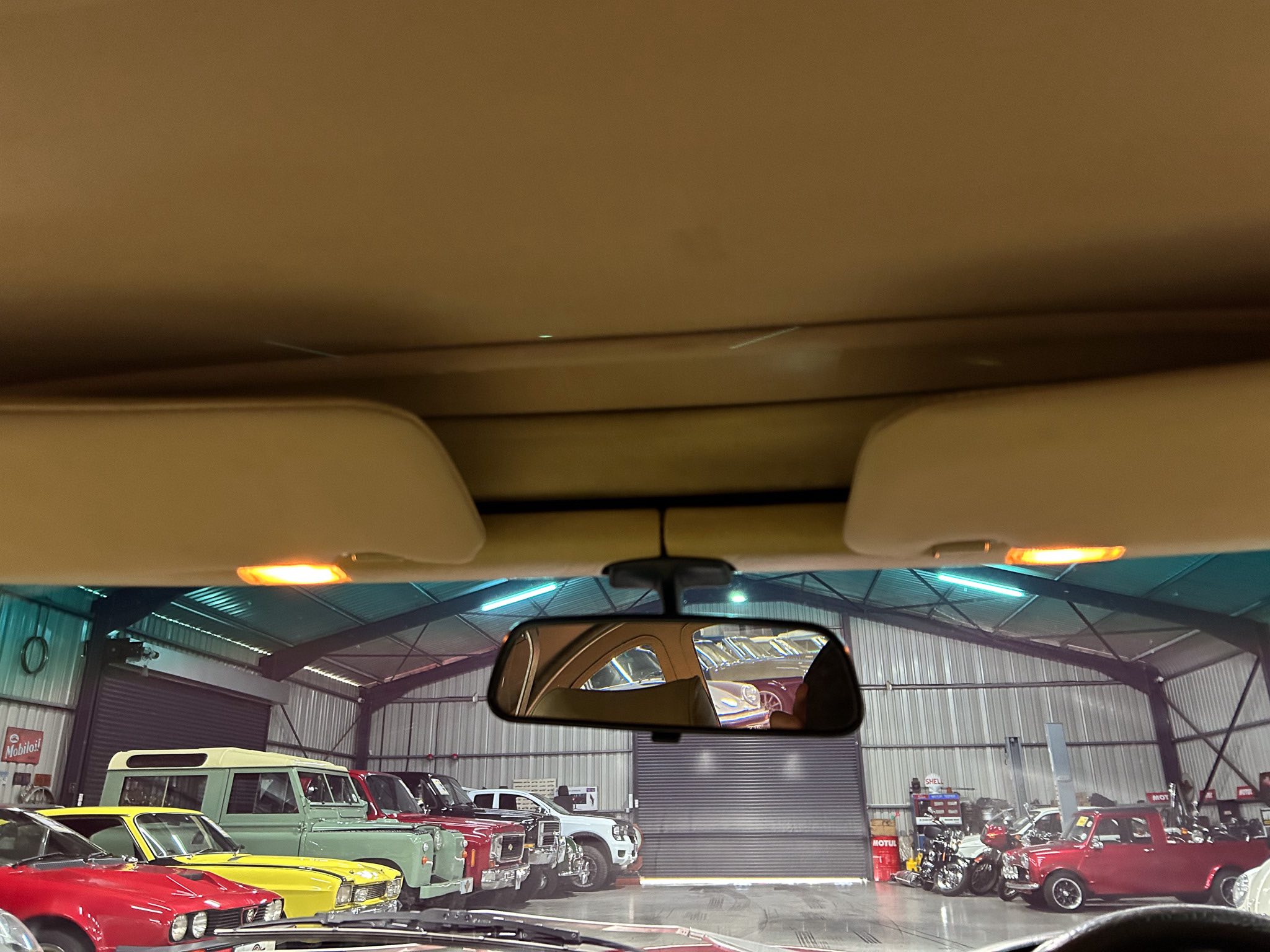 View from inside a car showroom: classic cars lined up under a high ceiling with orange lights, reflected in the rearview mirror.
