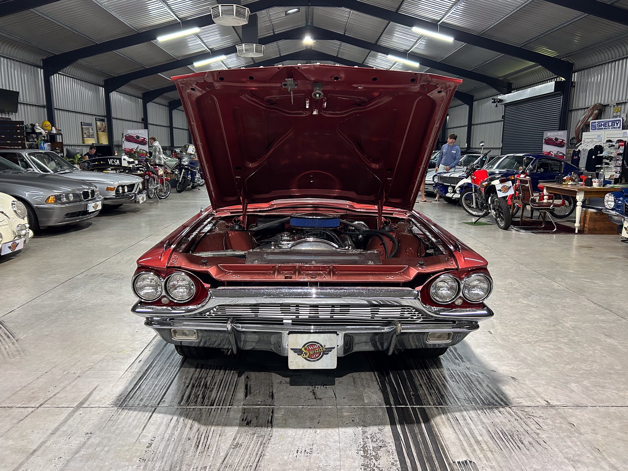 Red vintage car with its hood open in a showroom, exposing a blue air filter and engine bay, surrounded by other classic vehicles and motorcycles inside a metal-floored hangar.