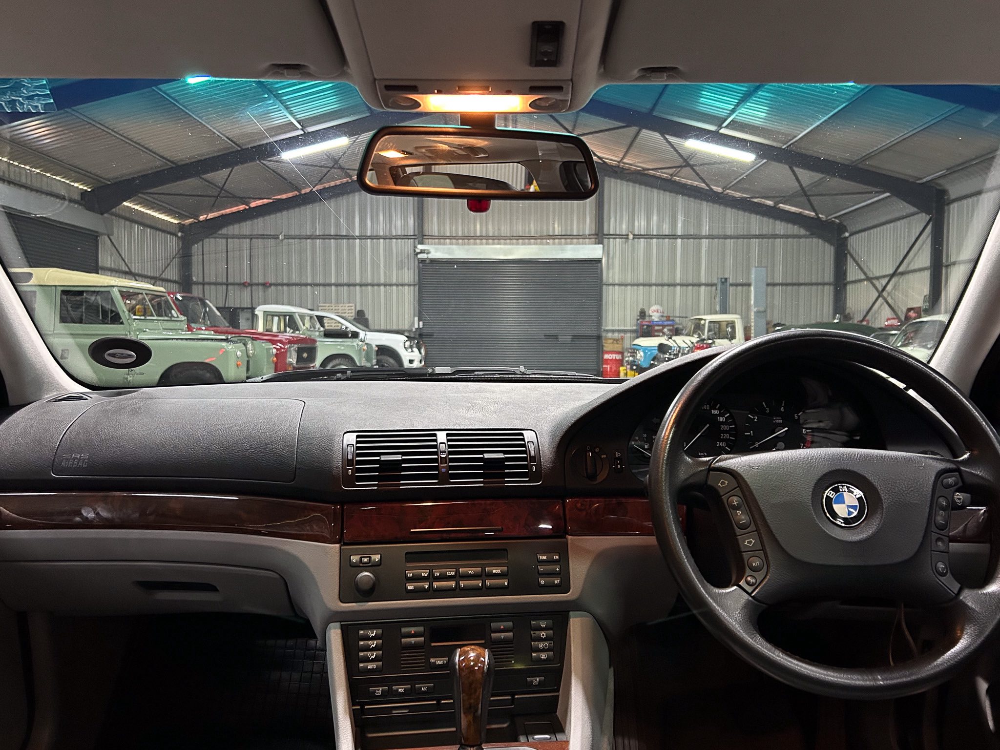 View of the car's dashboard and steering wheel from the driver's seat, looking through the windshield at a warehouse filled with vintage vehicles.