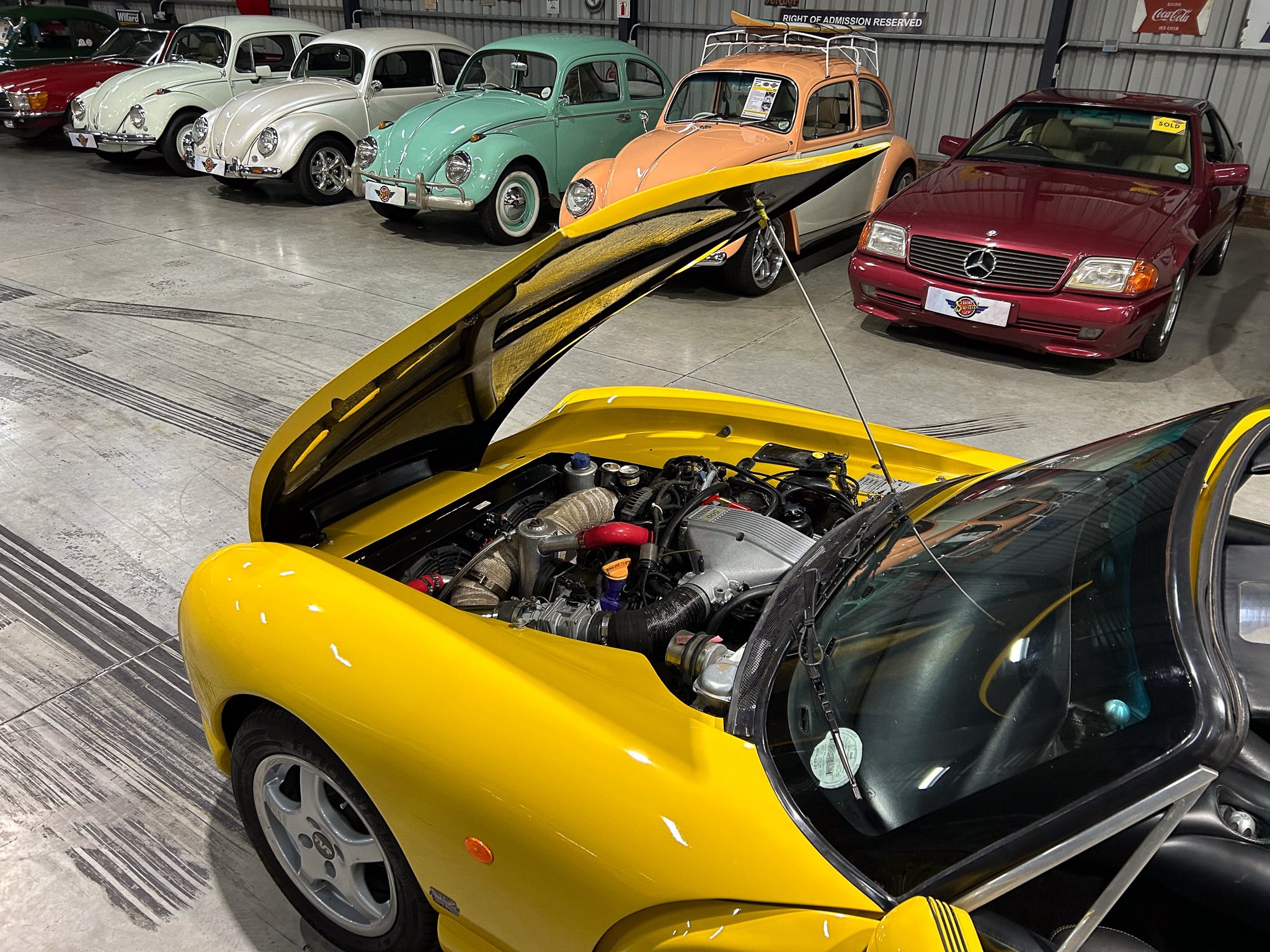 Row of vintage VW Beetles in pastel colors next to a maroon Mercedes inside a showroom, with a yellow sports car in the foreground having its hood open for inspection