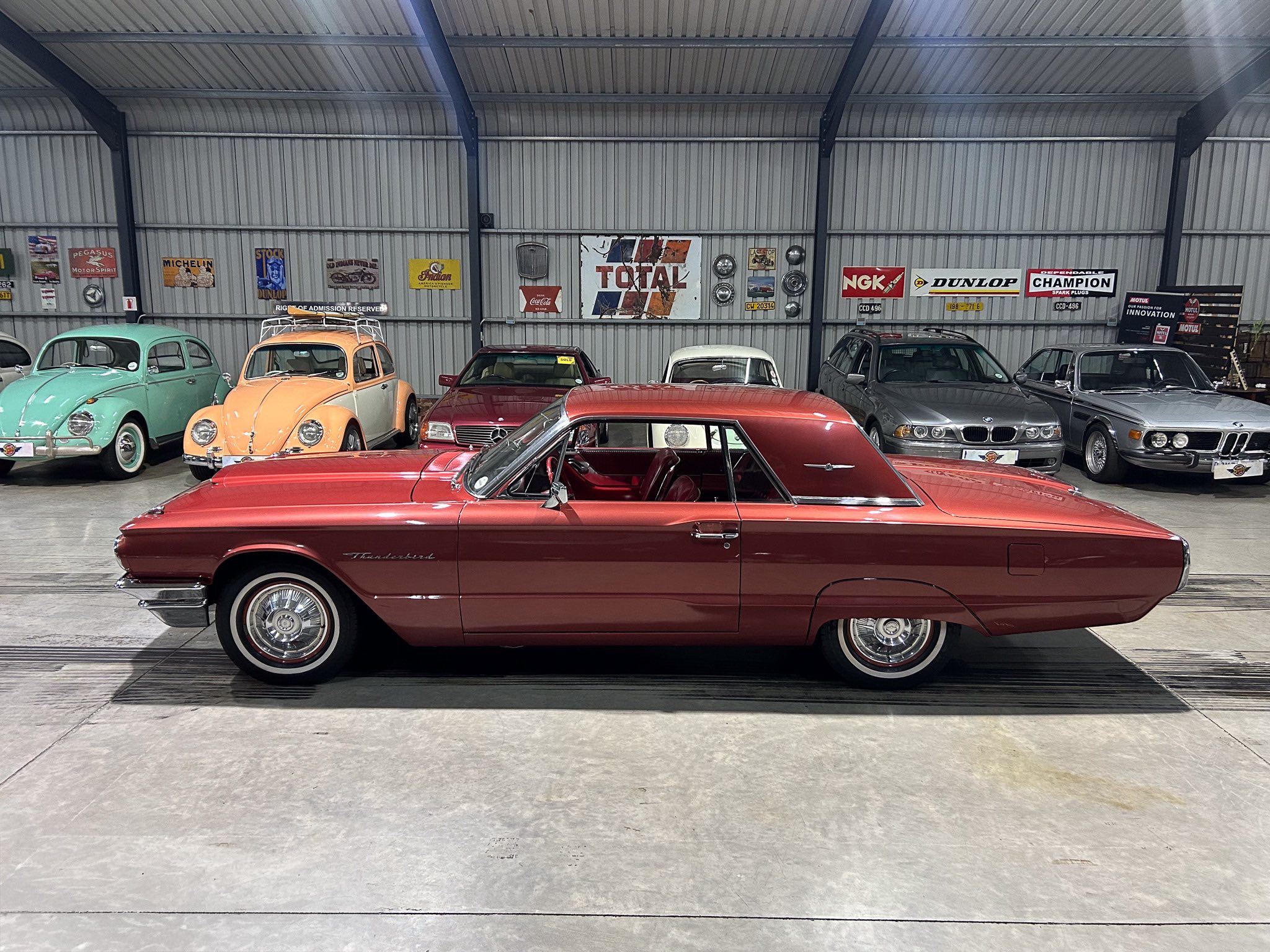Red Ford Thunderbird parked inside a vintage car showroom, with other classic cars against a wall of retro posters