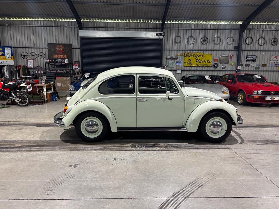 Side view of a cream-colored classic Volkswagen Beetle inside a car showroom with other vehicles and signs in the background