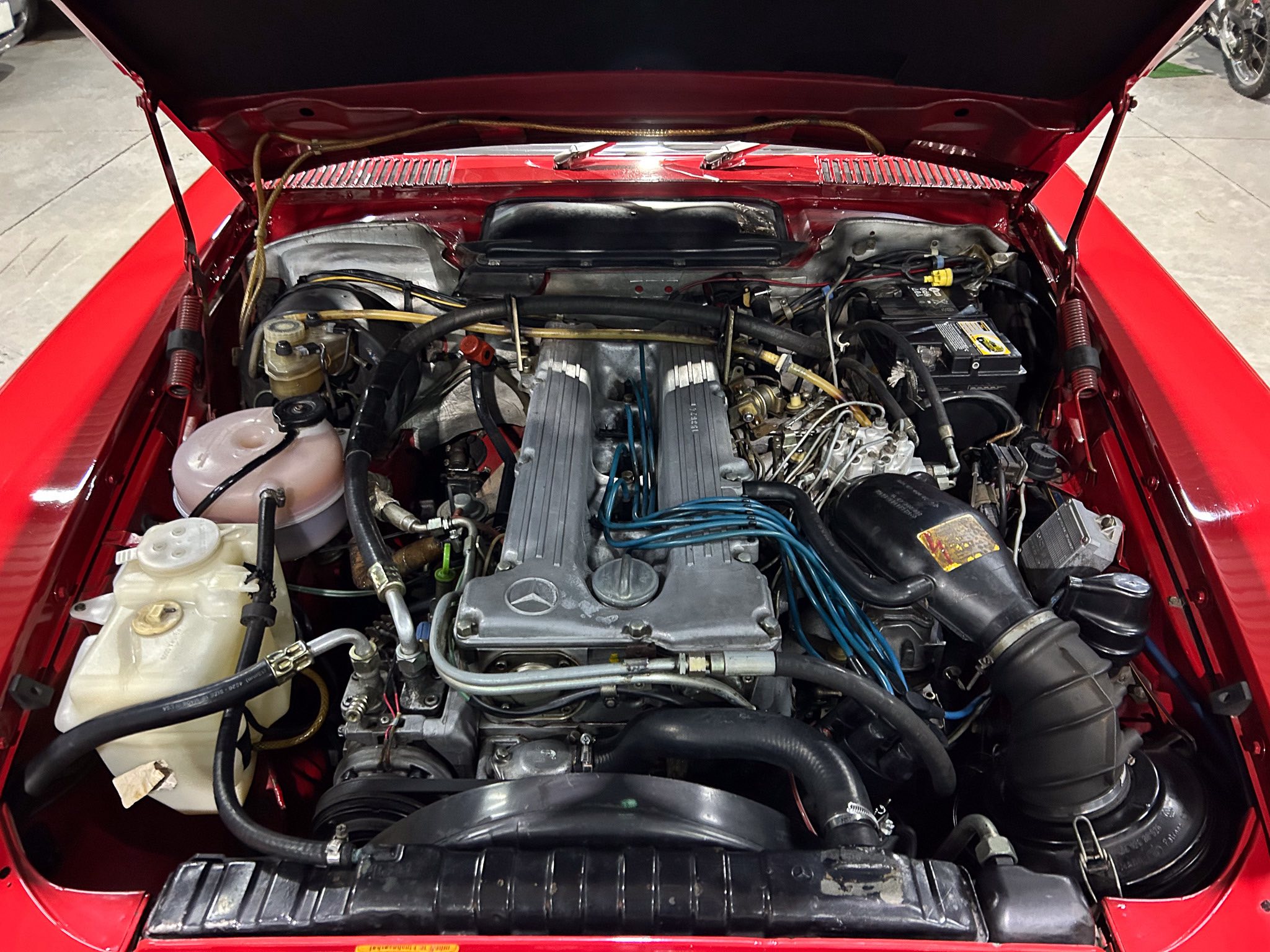 Open hood of a red classic car showing the engine bay with hoses, wires, and a battery on the right side