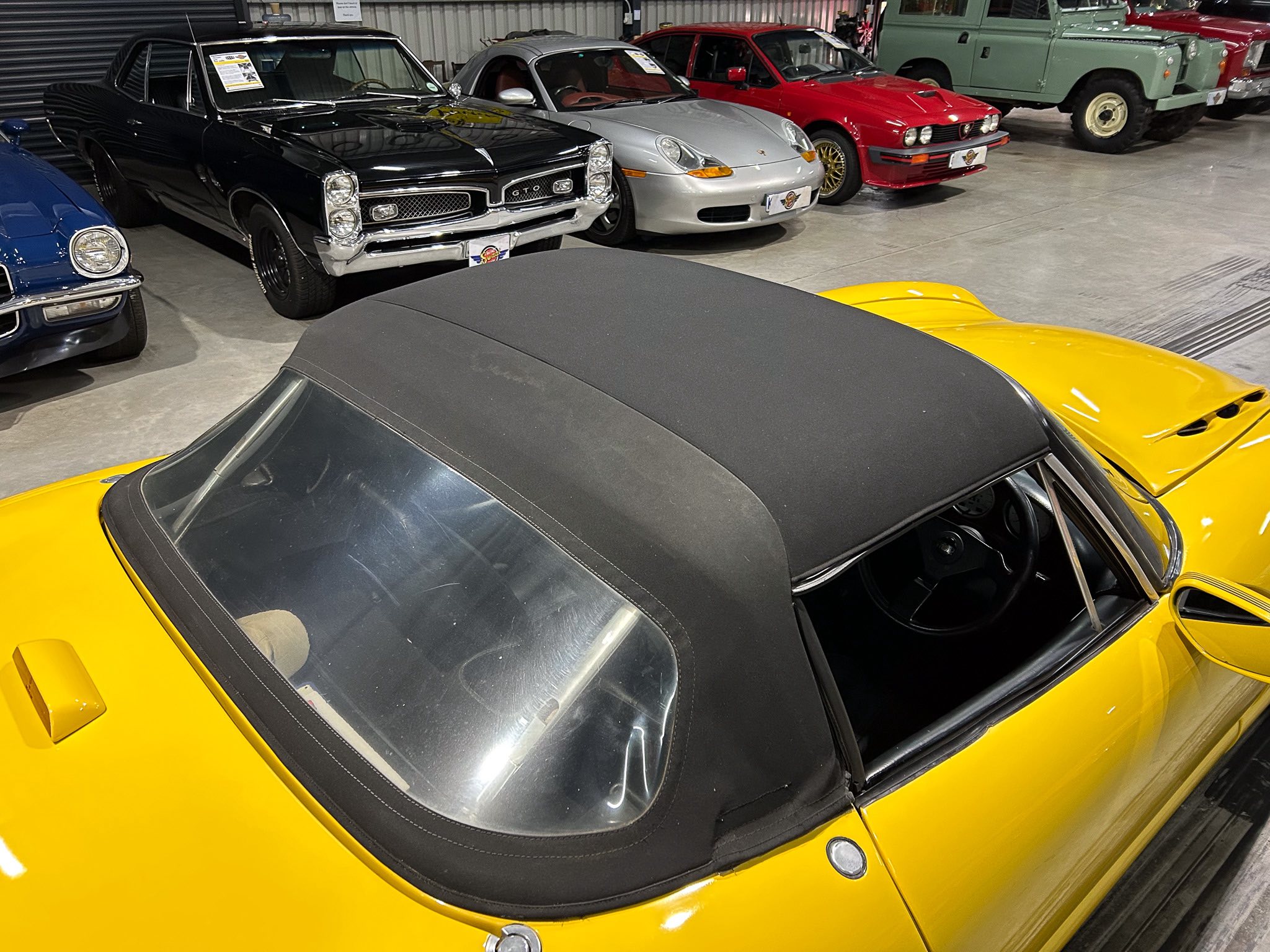 Close-up of a bright yellow convertible with a black soft top, parked among classic cars in a showroom.