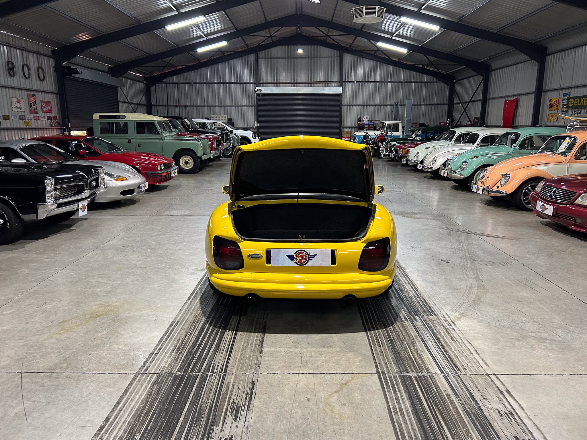 Bright yellow car with its trunk open, photographed inside a classic car showroom flanked by vintage vehicles on both sides.