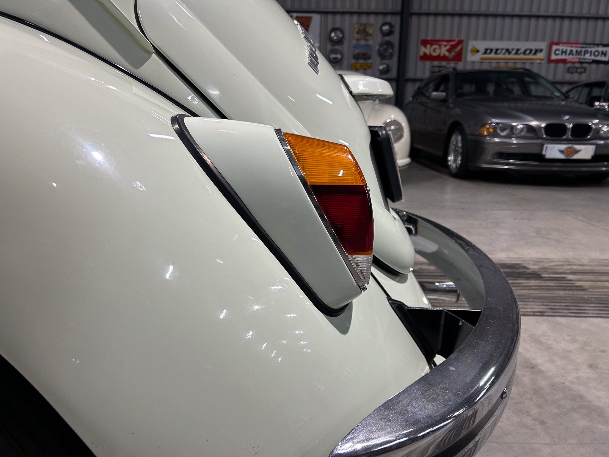 Close-up of a white vintage car’s rear end with an orange tail light and chrome bumper, showroom background with other cars and banners.