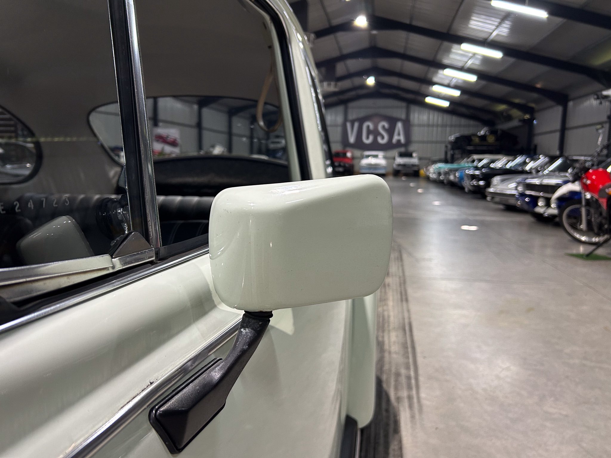 Close-up of a vintage car's side mirror and window trim inside a bright showroom with other classic cars in the background.