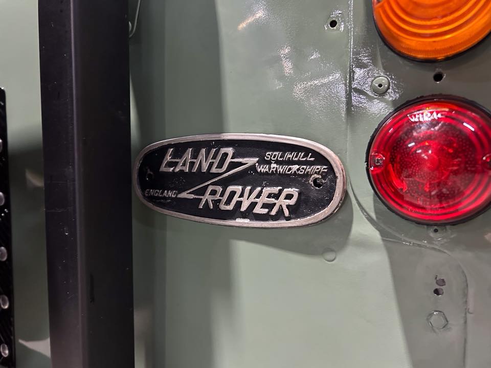 Close-up of a pale green vehicle showing a silver oval Land Rover badge beside a red tail light.