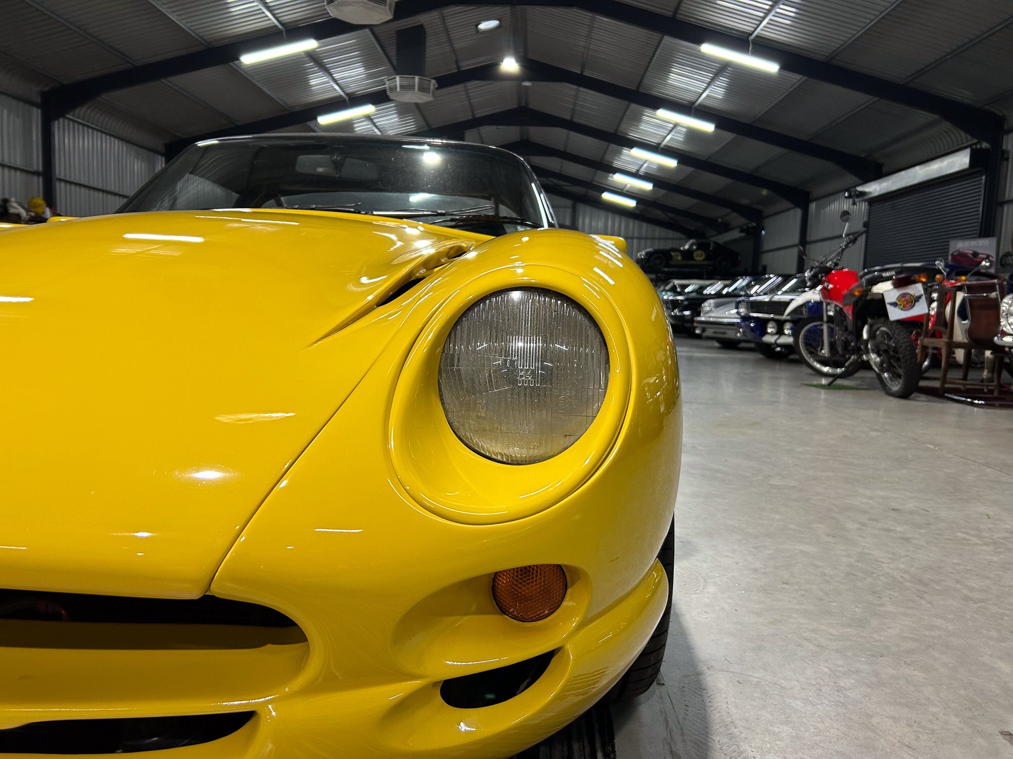 Close-up of a bright yellow vintage car's headlight and front bumper inside a workshop or showroom with other vehicles in the background.