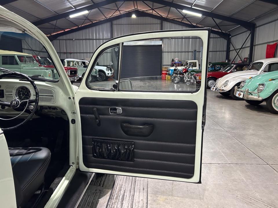 Open light-colored vintage car door showing black interior panel, with classic cars in a showroom behind.