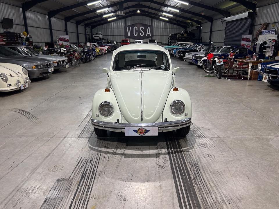 White vintage Volkswagen Beetle centered in a spacious showroom filled with classic cars in the background.