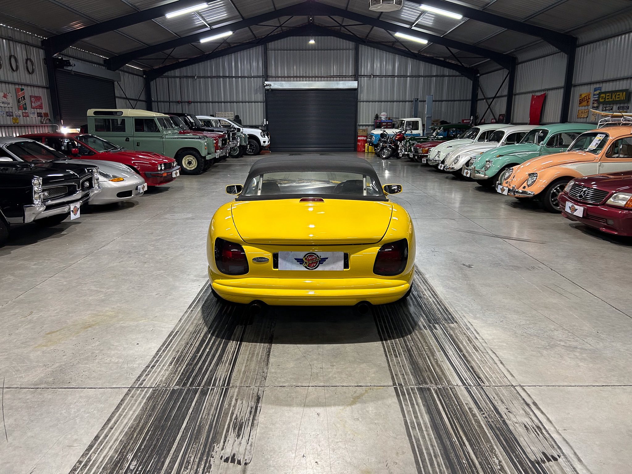 Bright yellow convertible viewed from behind, parked in a spacious showroom with many vintage cars along the sides.