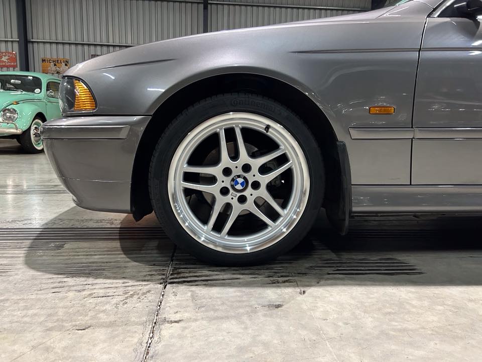 Close-up of a silver BMW car's front wheel and fender, showing a multi-spoke alloy rim and tire in a showroom.
