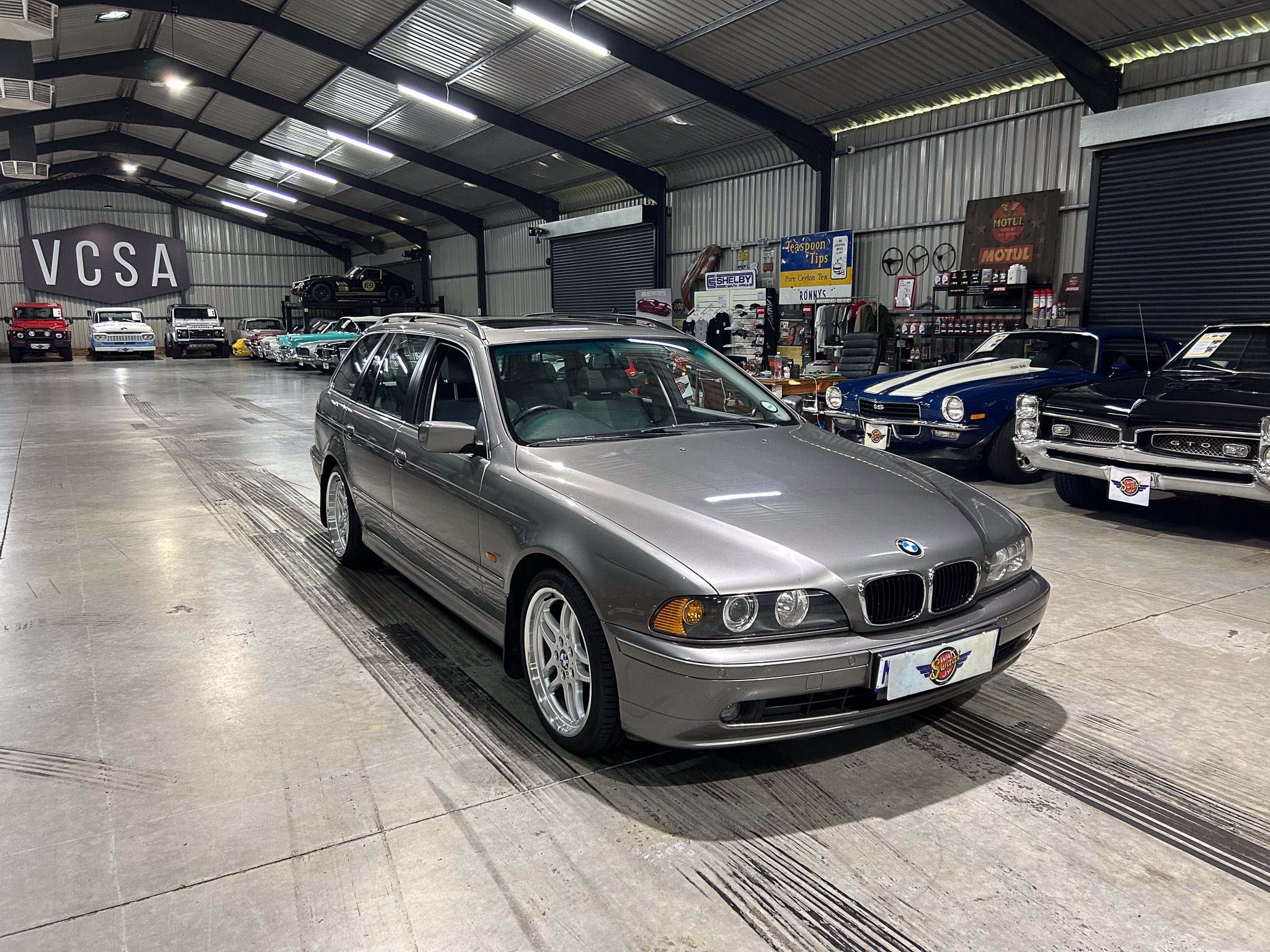 Inside a large car showroom with a row of classic and modern cars; a silver BMW in the foreground and vintage vehicles along the left wall under a VCSA sign.