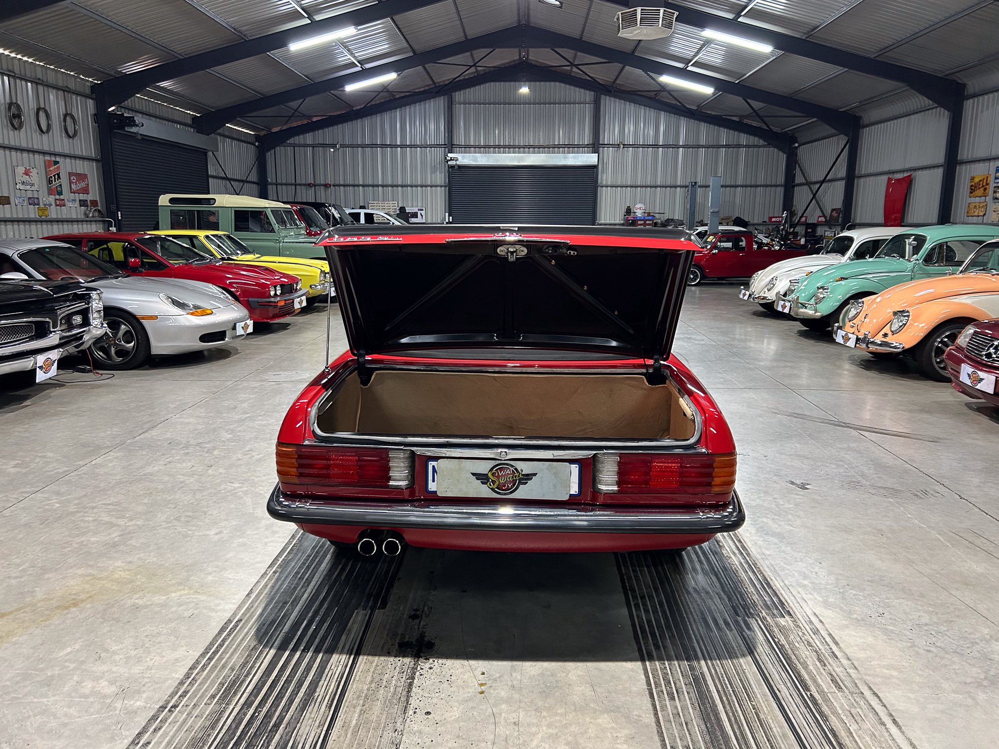 Rear view of a red classic car with its trunk open in a large showroom filled with vintage vehicles on both sides.