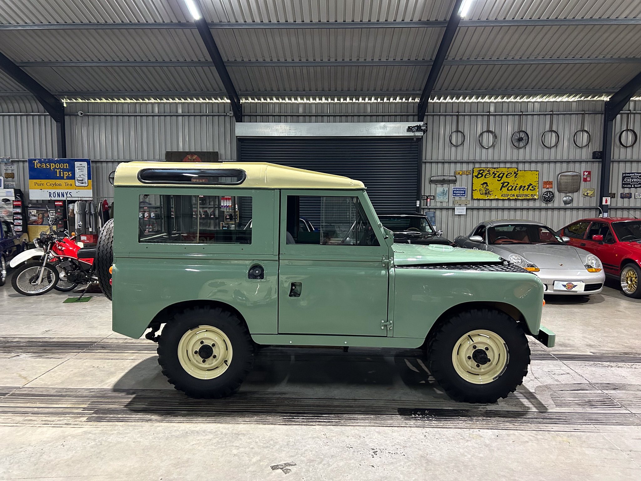 Inside a metal workshop showing a vintage light-green utility vehicle parked among bikes and retro signs on the wall.