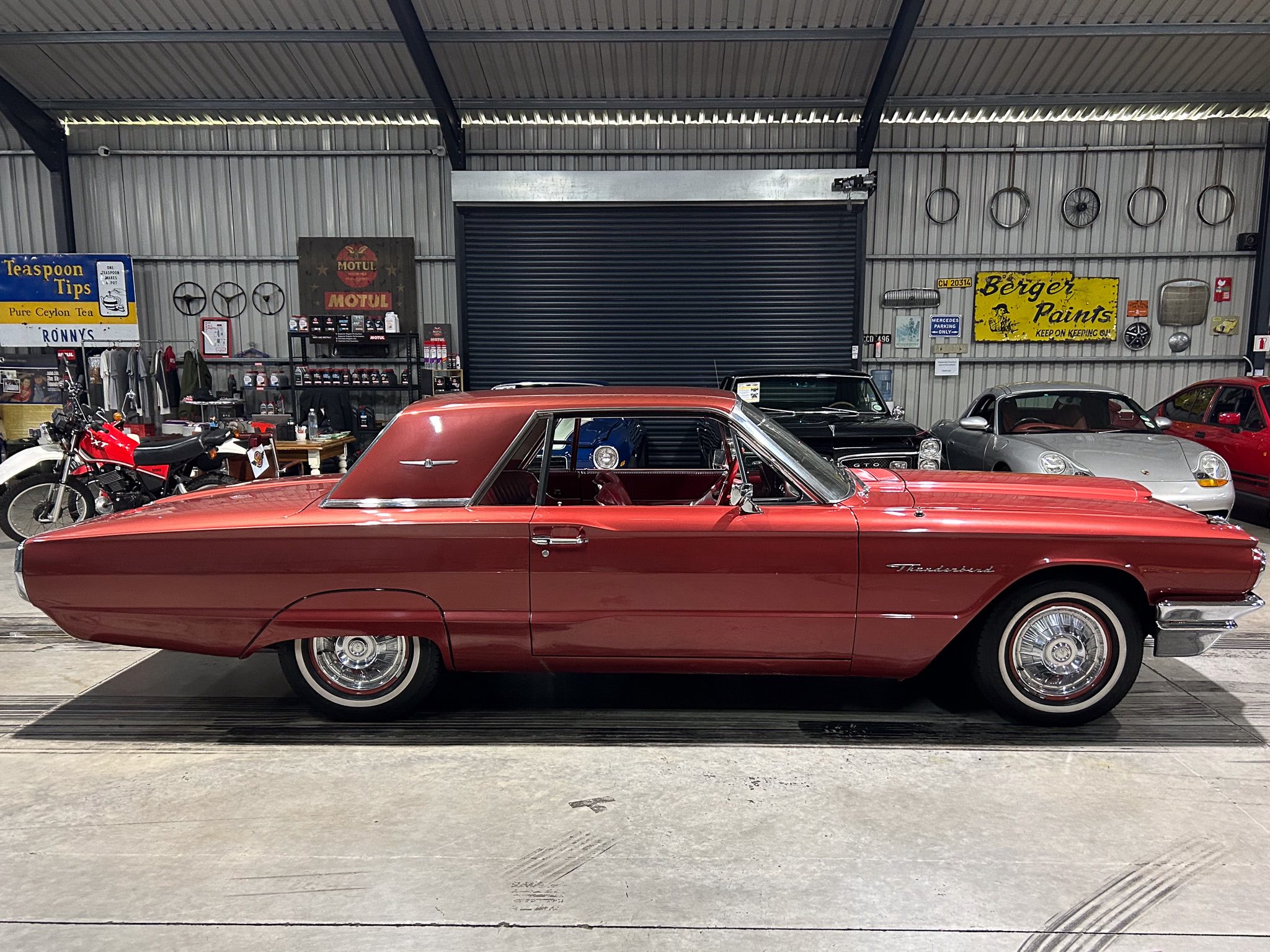 Red vintage car in a garage showroom with chrome trim, foreground dominating the scene and retro signs on the wall in the background to the left and right.