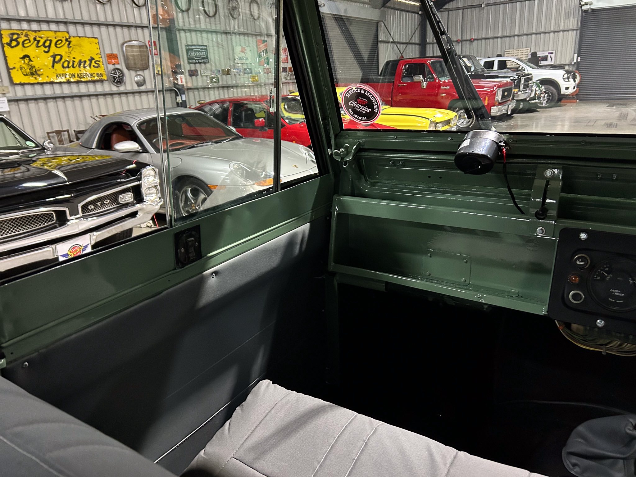 Interior of a green vintage truck cab with a visible dashboard, windows revealing a garage full of classic cars and a yellow 'Berger Paints' sign on the wall