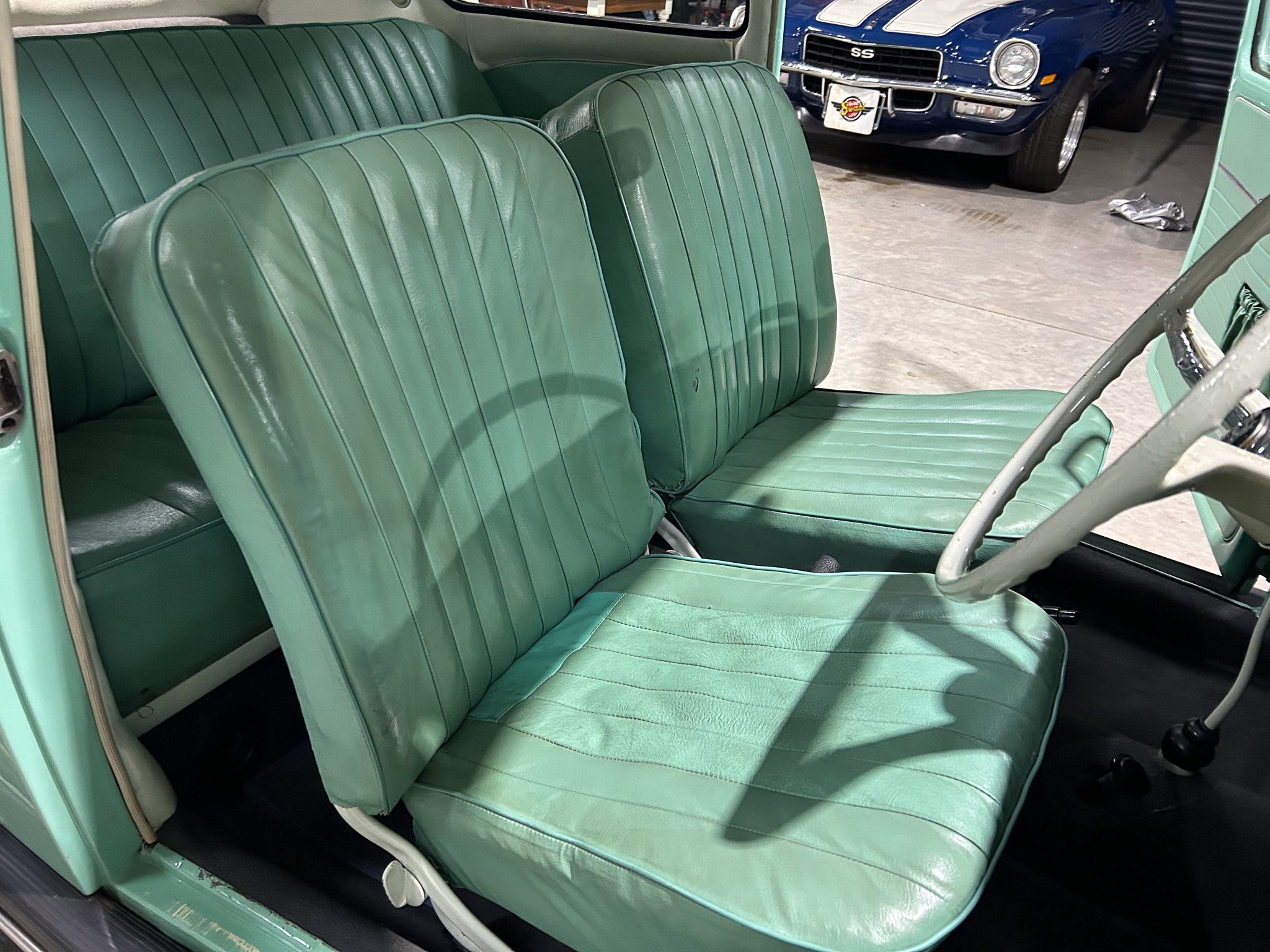 Vintage car interior with teal-green, pleated leather seats in foreground and a blue classic car visible through the open door in the background