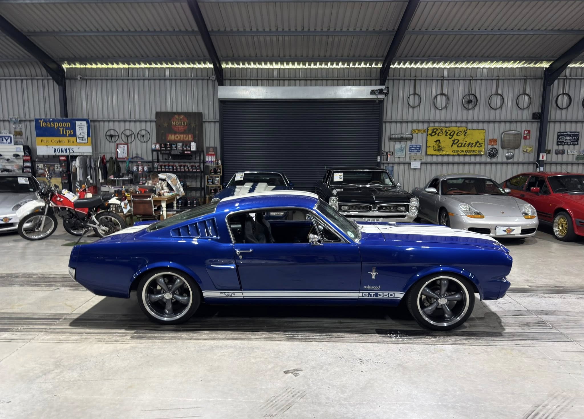 Blue Ford Mustang GT350 in a classic car showroom, surrounded by other cars and bikes.