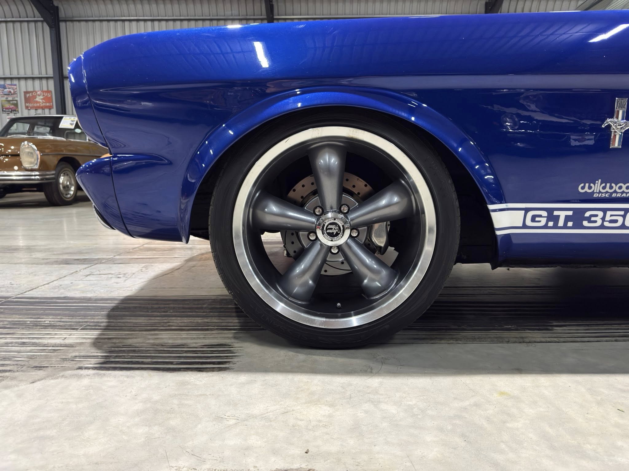 Close-up of a blue car's wheel and fender, with a polished five-spoke rim and brake disc visible inside a showroom-like space.