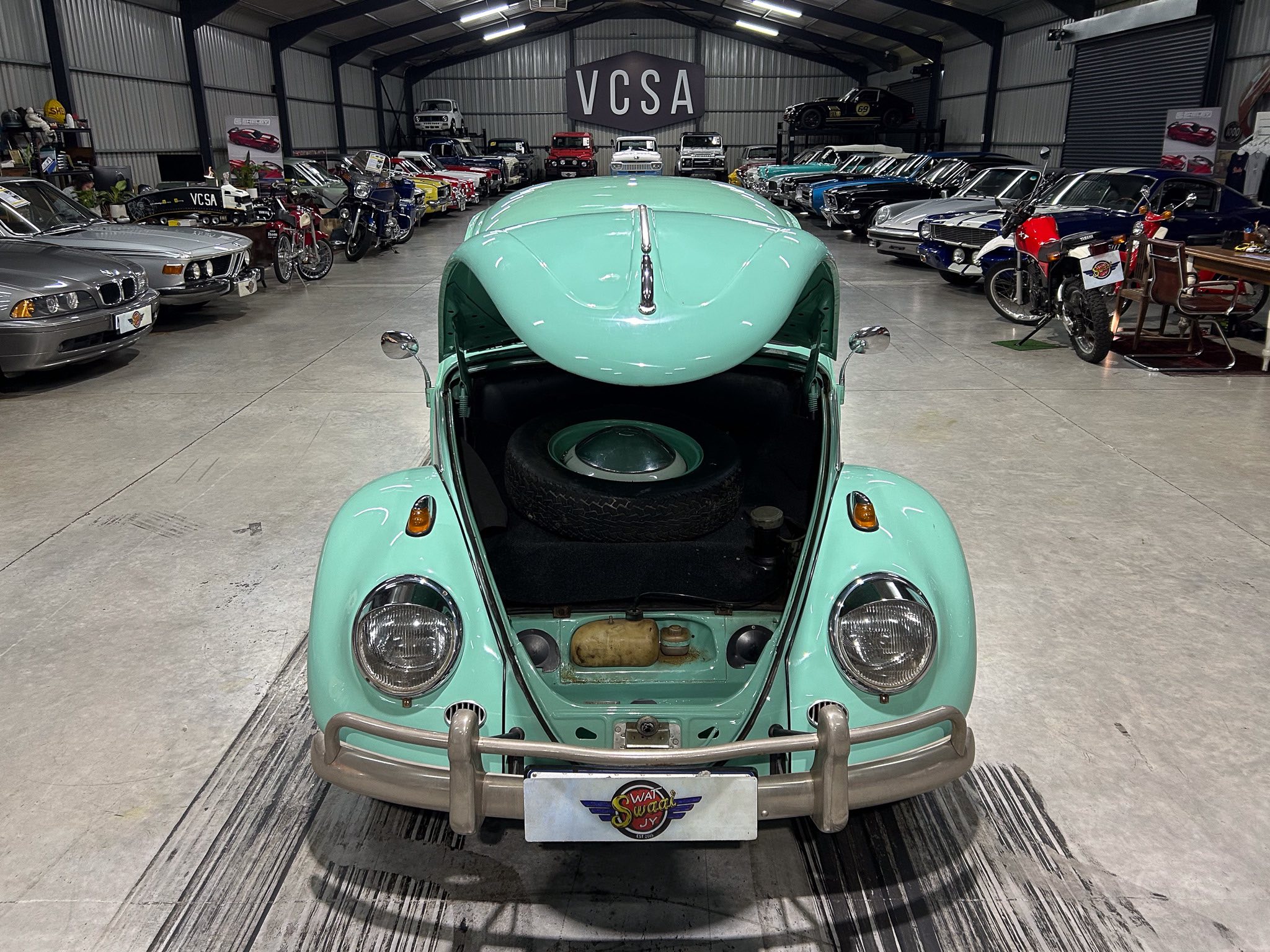 Mint-green vintage car from the rear, parked inside a large showroom filled with classic cars lined on both sides and a VCSA sign overhead.