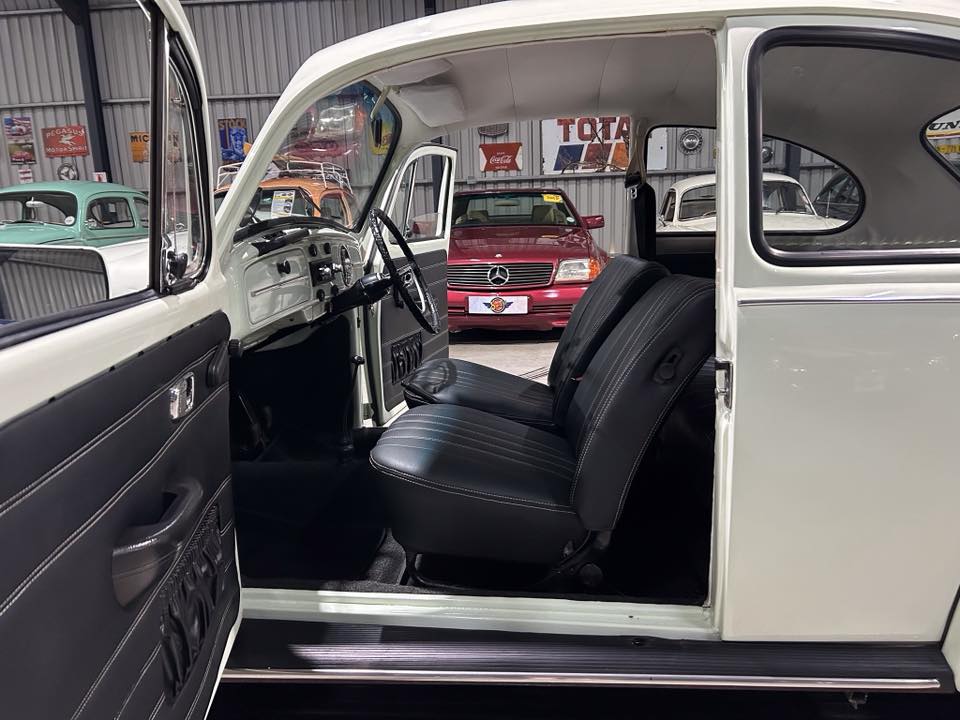 Interior of a vintage white car with the driver's door open, showing black leather seats and a white dashboard inside a showroom with classic cars in the background