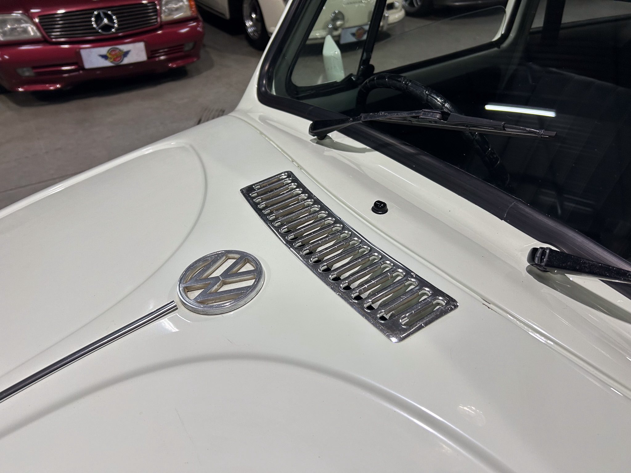 Close-up of a white car hood with a circular VW emblem and chrome vents, windshield wipers resting on the glass in a showroom garage.