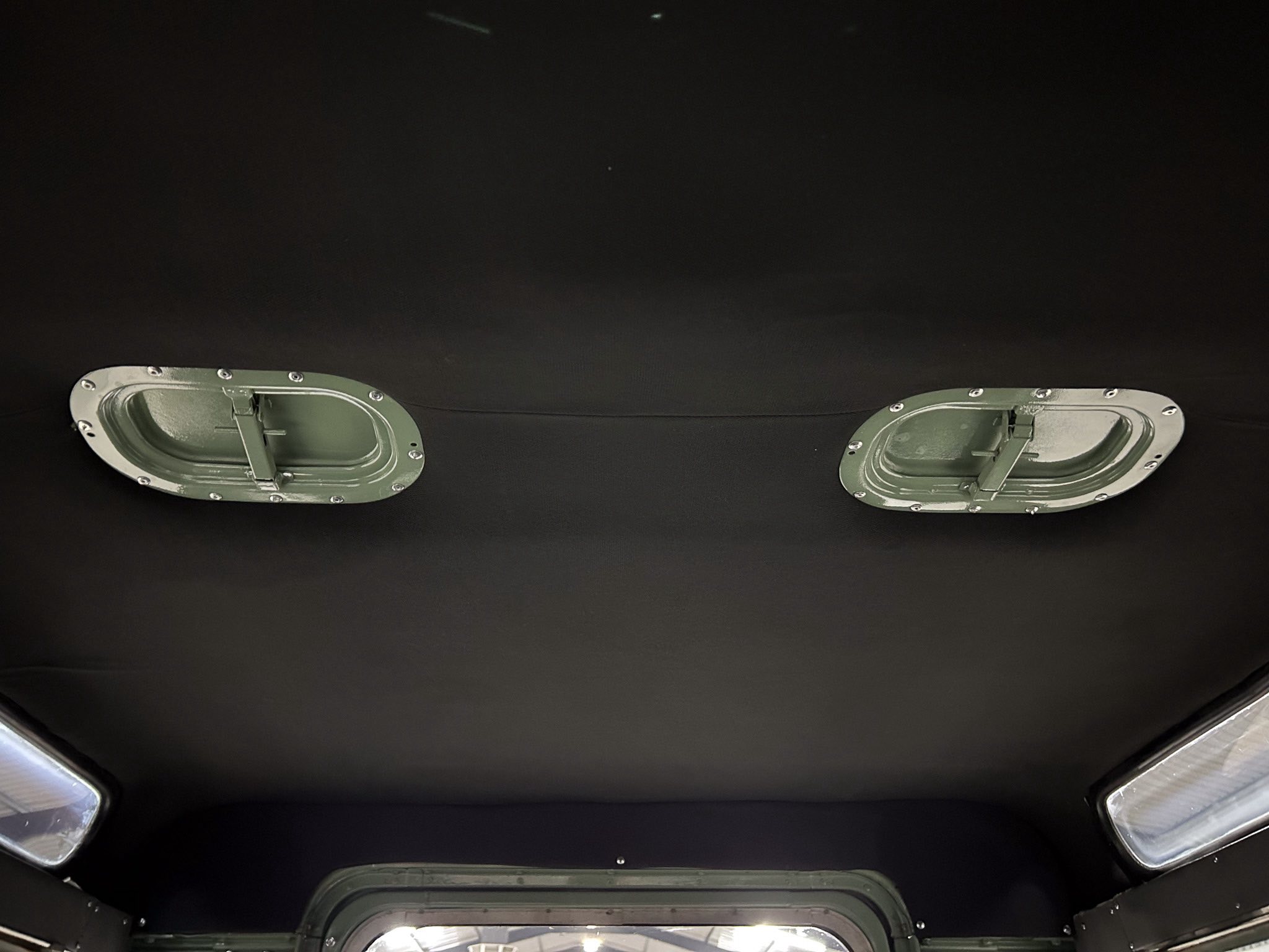 Dark vehicle interior ceiling featuring two oval metal overhead hatches with bolted rims along the top edge of the image.