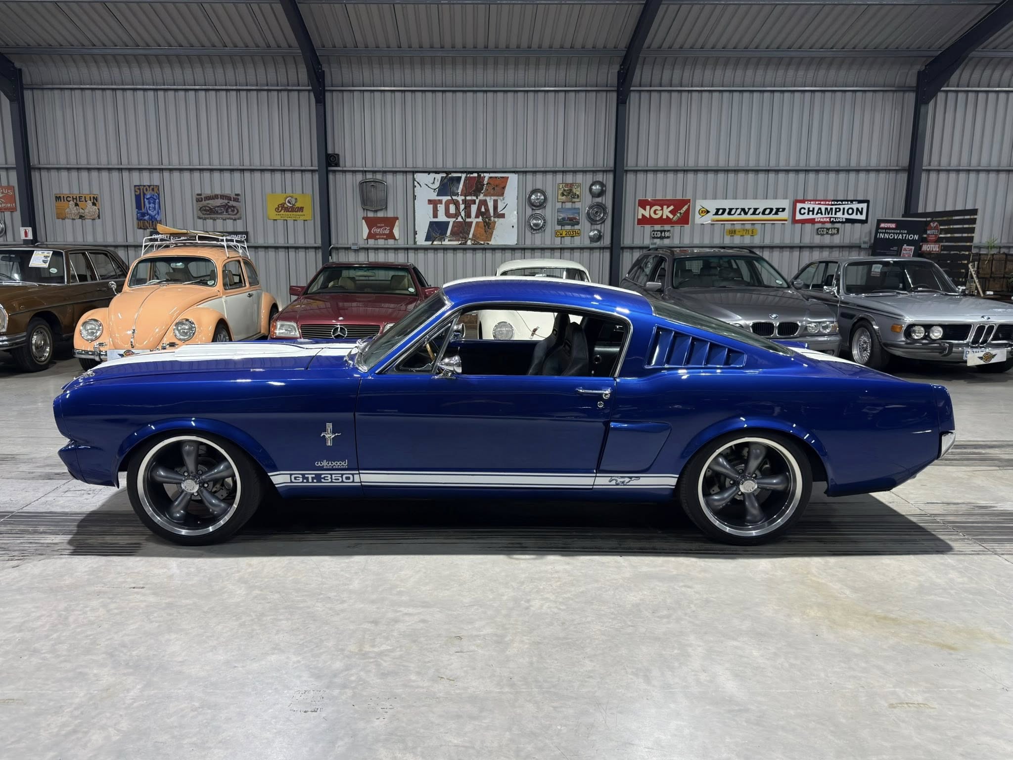 Blue 1965 Ford Mustang GT350 in a showroom, with other vintage cars and retro signs on a metal wall.