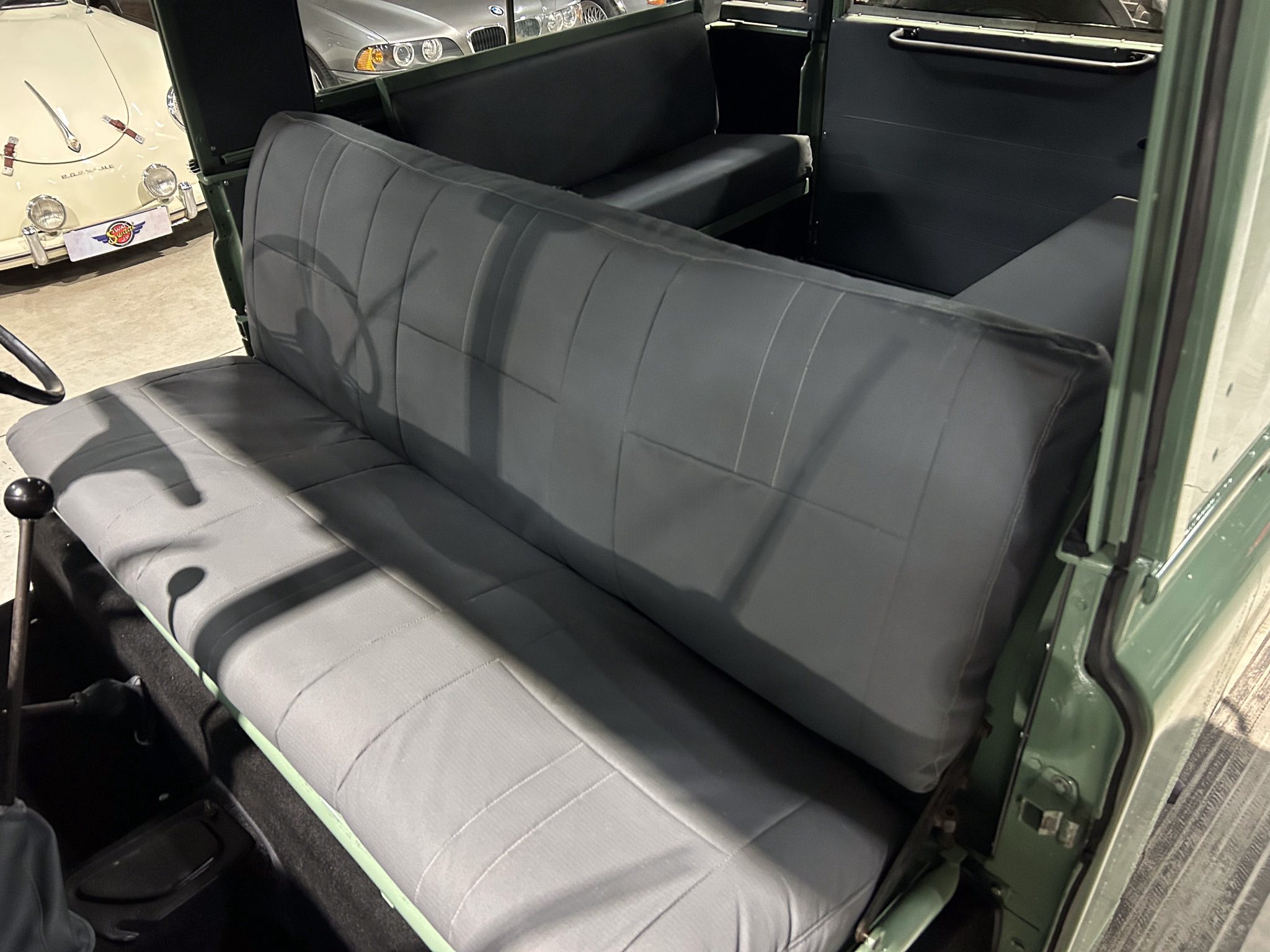 Interior of a vintage green car showing gray bench seats and door panel; other classic cars visible through the window.