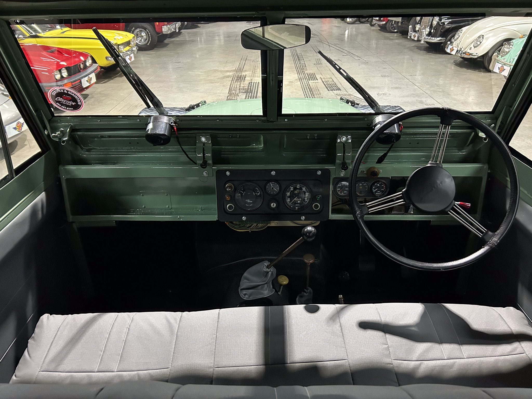 Front cab view of a vintage green vehicle, showing dashboard, large steering wheel, windshield wipers, and a showroom full of classic cars outside the windshield.
