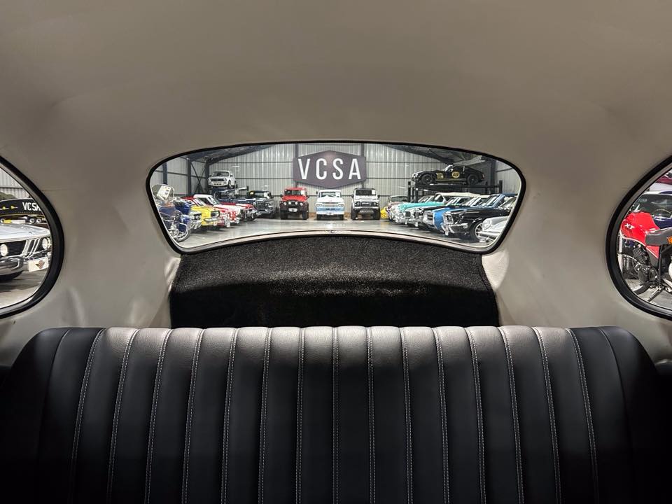 Backseat view of a vintage car, with a showroom of colorful classic cars visible through the rear window.